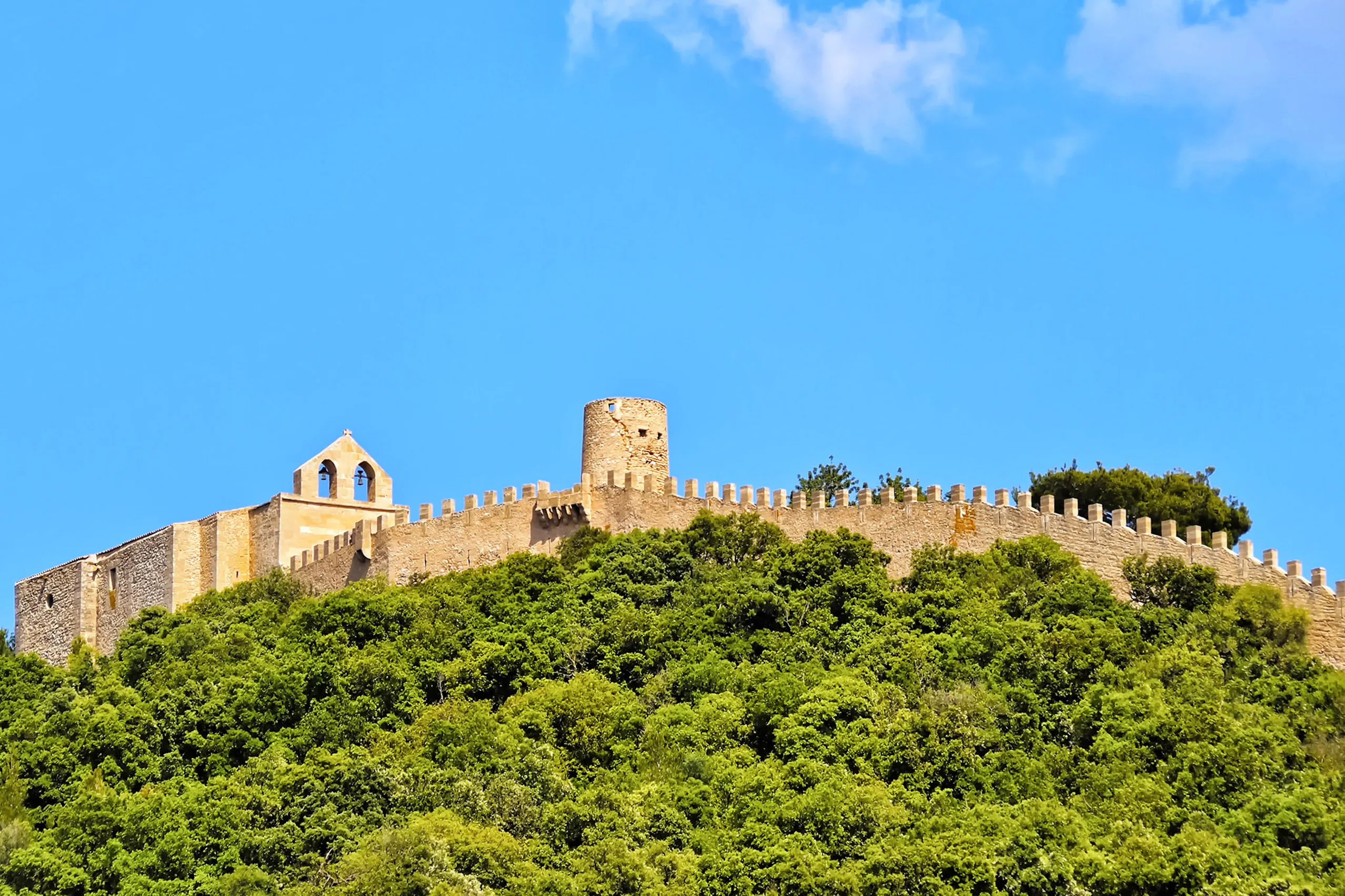 Das Castell de Capdepera auf einem dicht bewachsenen Hügel: eine lange Zinnenmauer mit Wehrturm und einem Gebäude mit Glockengiebel vor blauem Himmel.
