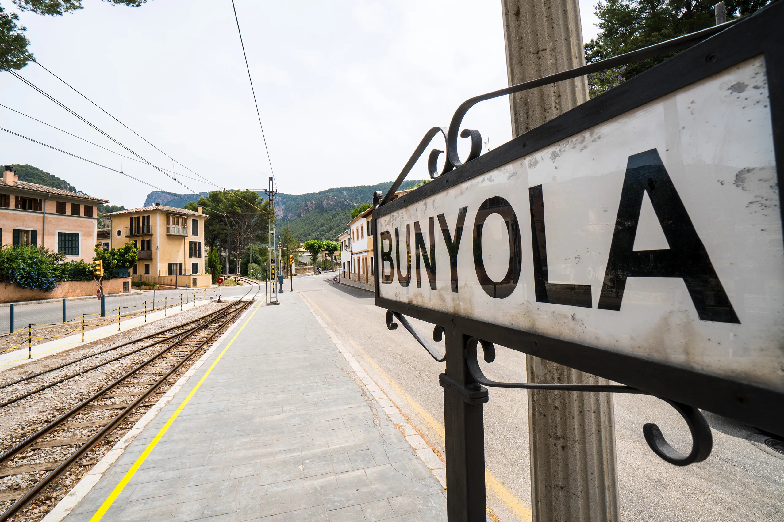 Weißes emailliertes Bahnhofsschild mit der Aufschrift BUNYOLA an einem geschwungenen Halter über dem Gleis, dahinter Bahnsteig und Berge.
