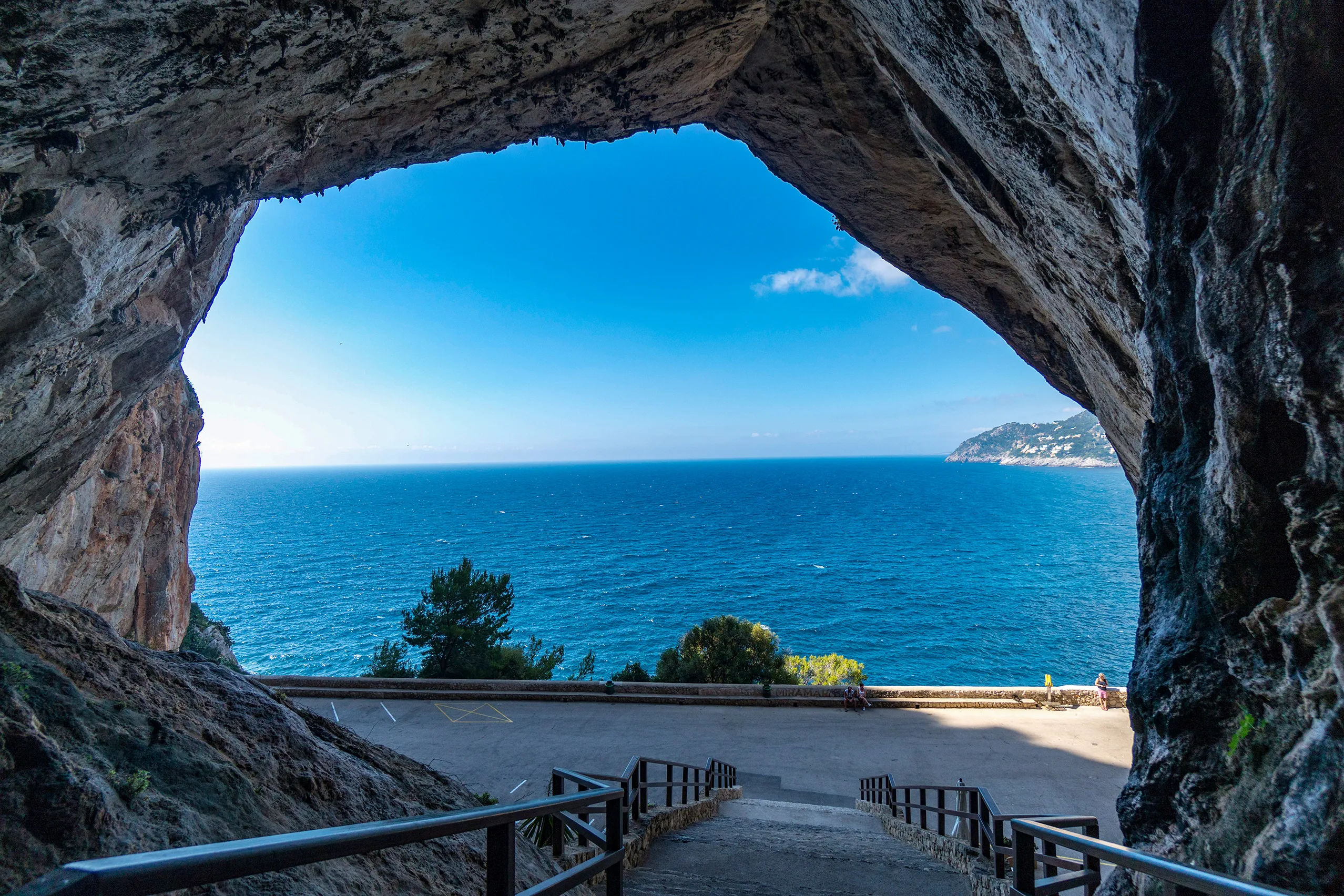 Blick aus dem hohen, dunklen Portal der Coves d'Artà hinaus auf das blaue Meer und die bewaldete Küste, im Vordergrund ein Geländer und der Felsboden.
