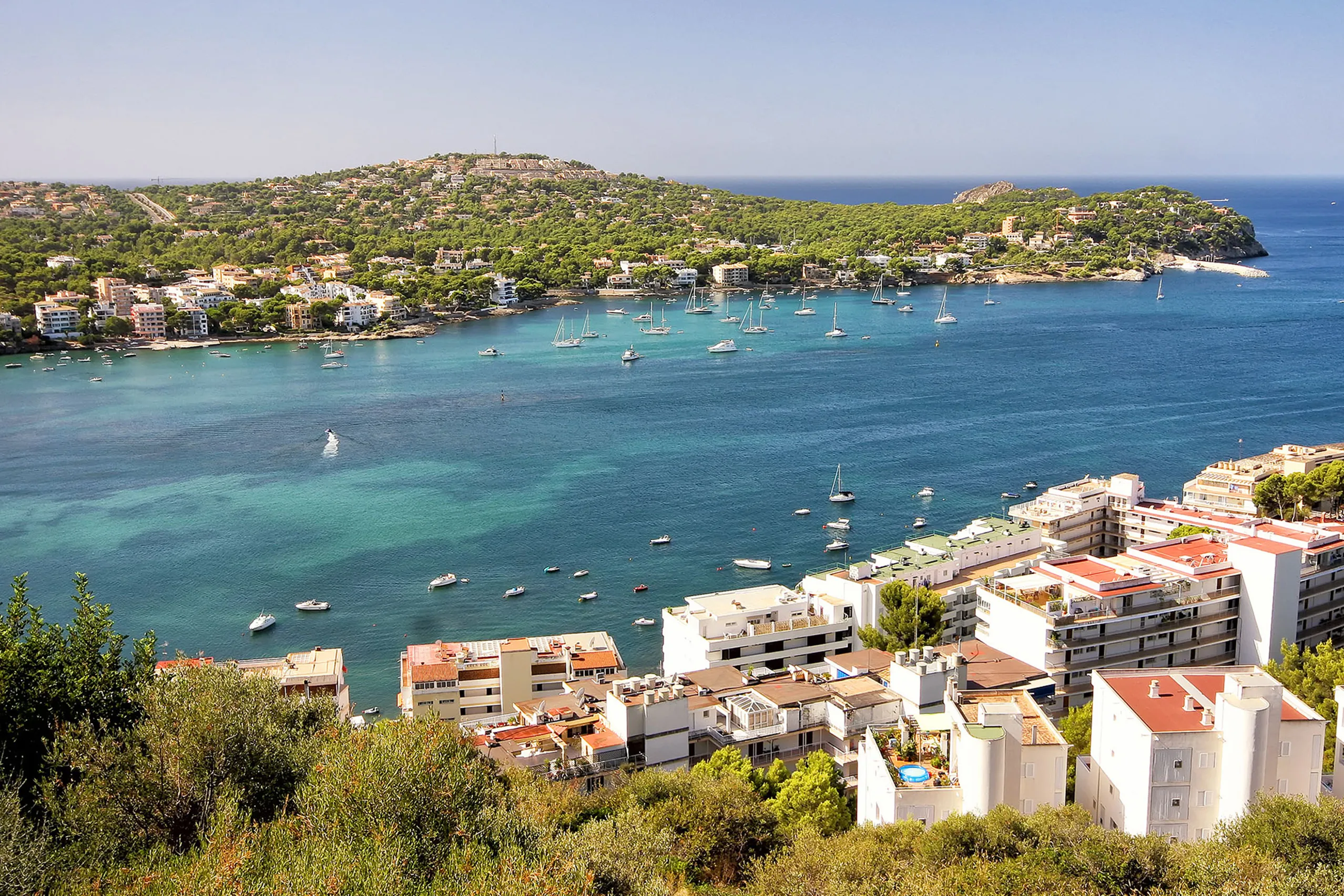 Blick von oben über die Bucht von Santa Ponça mit türkisem Wasser, ankernden Booten, der bewaldeten Landzunge und den weißen Häusern des Orts im Vordergrund.
