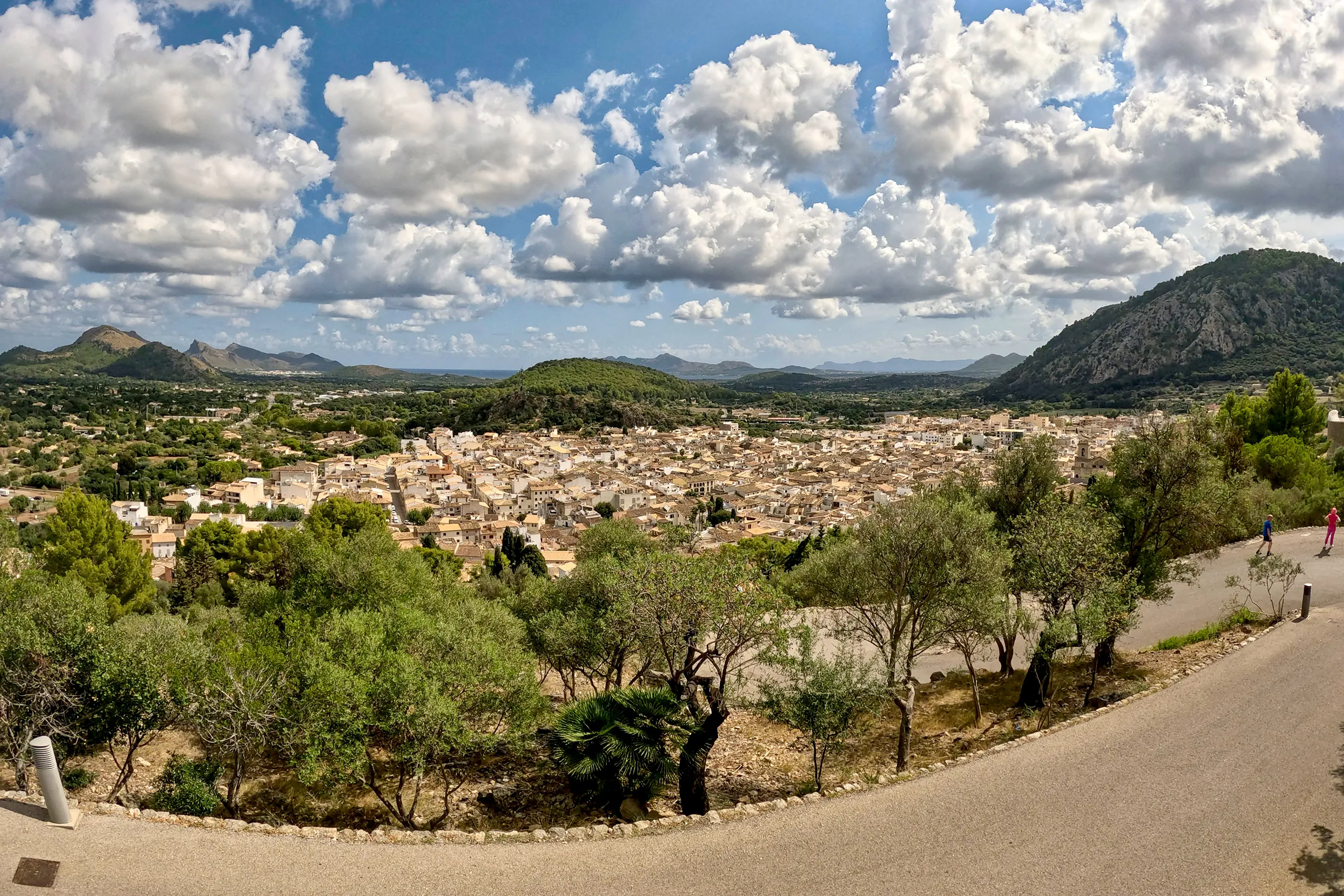 Blick von der Straße zum Kalvarienberg über die dicht gedrängten Ziegeldächer von Pollença; links und rechts bewaldete Bergkuppen, dahinter die Ebene und in der Ferne das Meer unter einem Himmel voller Quellwolken.
