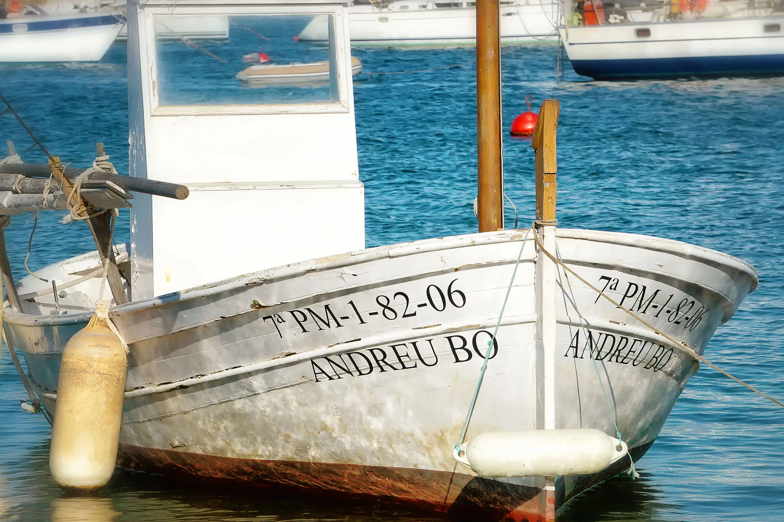 Reihe farbiger Bootsschuppen mit blauen, grünen und roten Toren am Ufer von Portocolom, davor liegende Fischerboote im ruhigen Wasser des Naturhafens.
