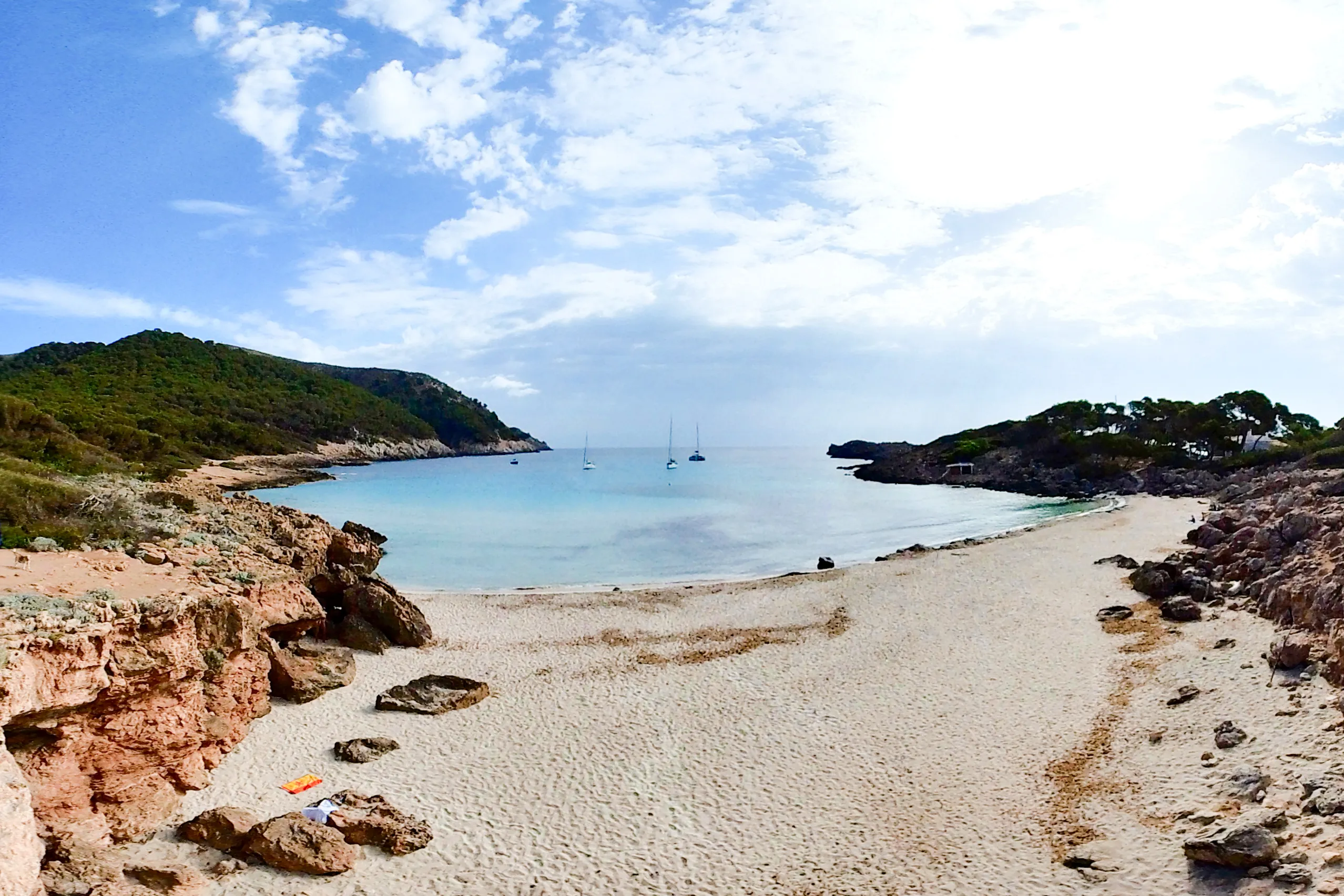 Kleine Bucht der Cala Moltó mit hellem Sandstreifen zwischen Felsen, im ruhigen Wasser ankern zwei Segelboote, dahinter eine bewaldete Landzunge.
