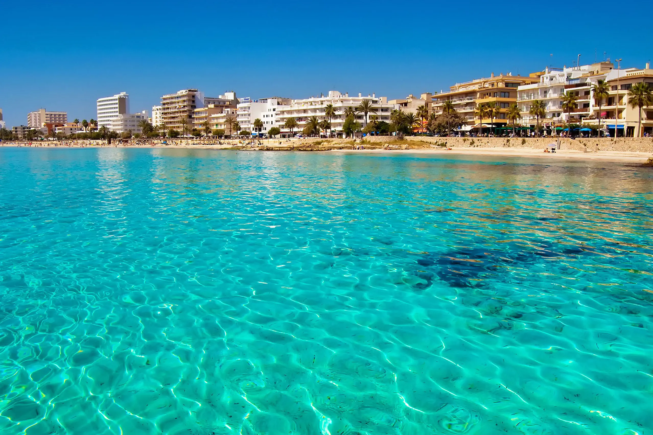 Die weite Bucht von Cala Millor mit türkisfarbenem klarem Wasser, hellem Sandstrand und der Hotelzeile dahinter unter blauem Himmel.
