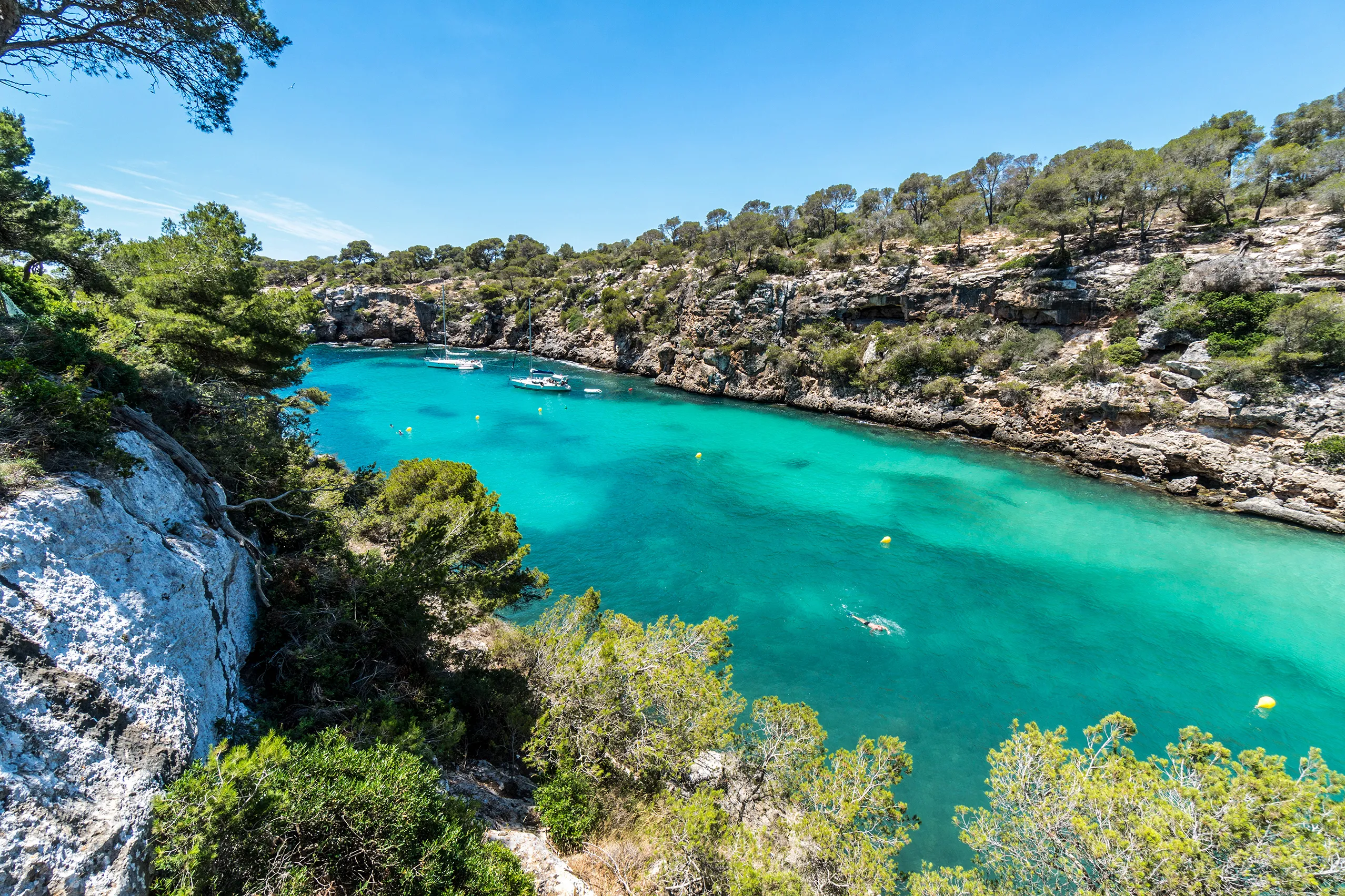 Blick von der Klippe in die schmale Bucht von Cala Pi mit türkisgrünem Wasser zwischen hellen, dicht bewachsenen Felswänden, im Hintergrund das offene Meer.
