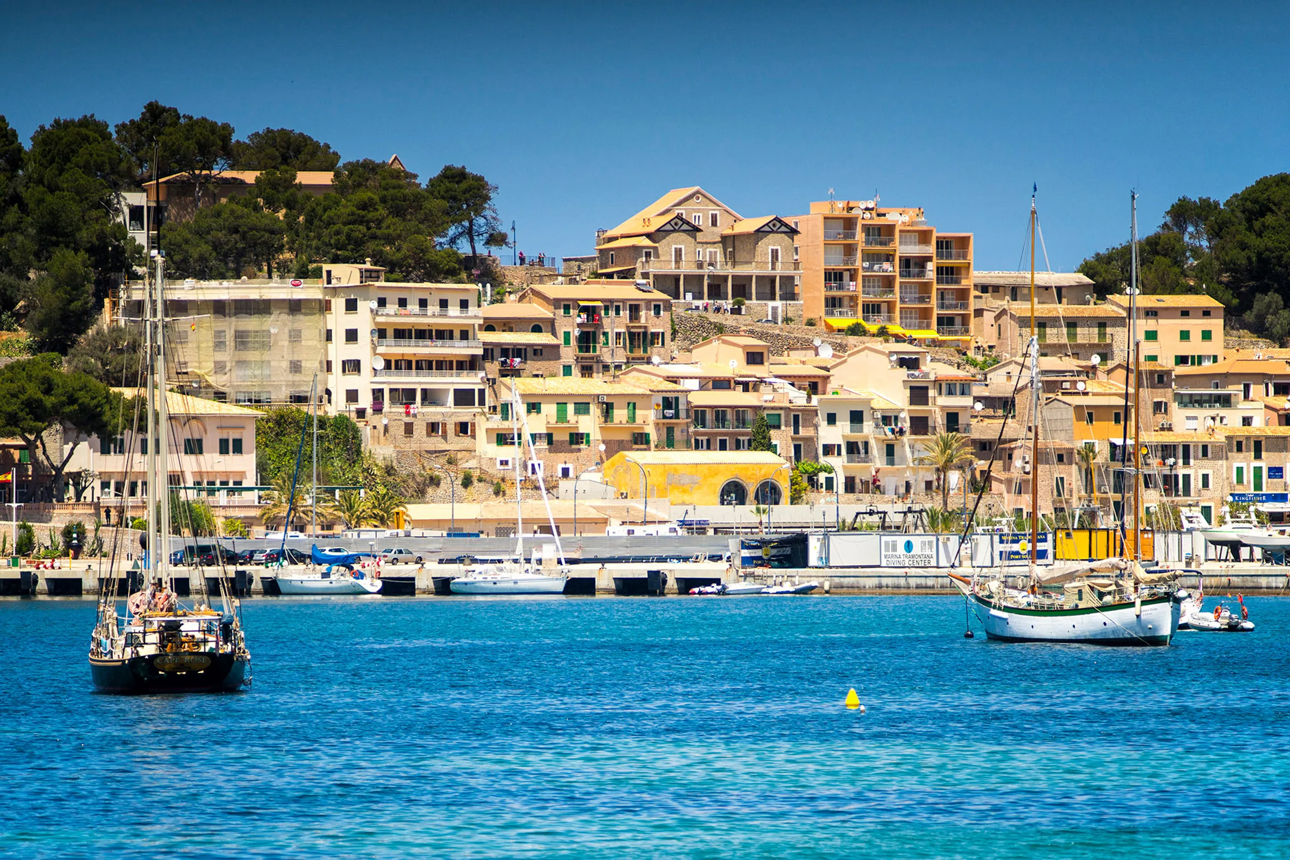 Blick von oben auf die fast kreisrunde Bucht von Port de Sóller mit Yachthafen, Häusern am Ufer, bewaldeten Hängen und der schmalen Einfahrt zum offenen Meer.
