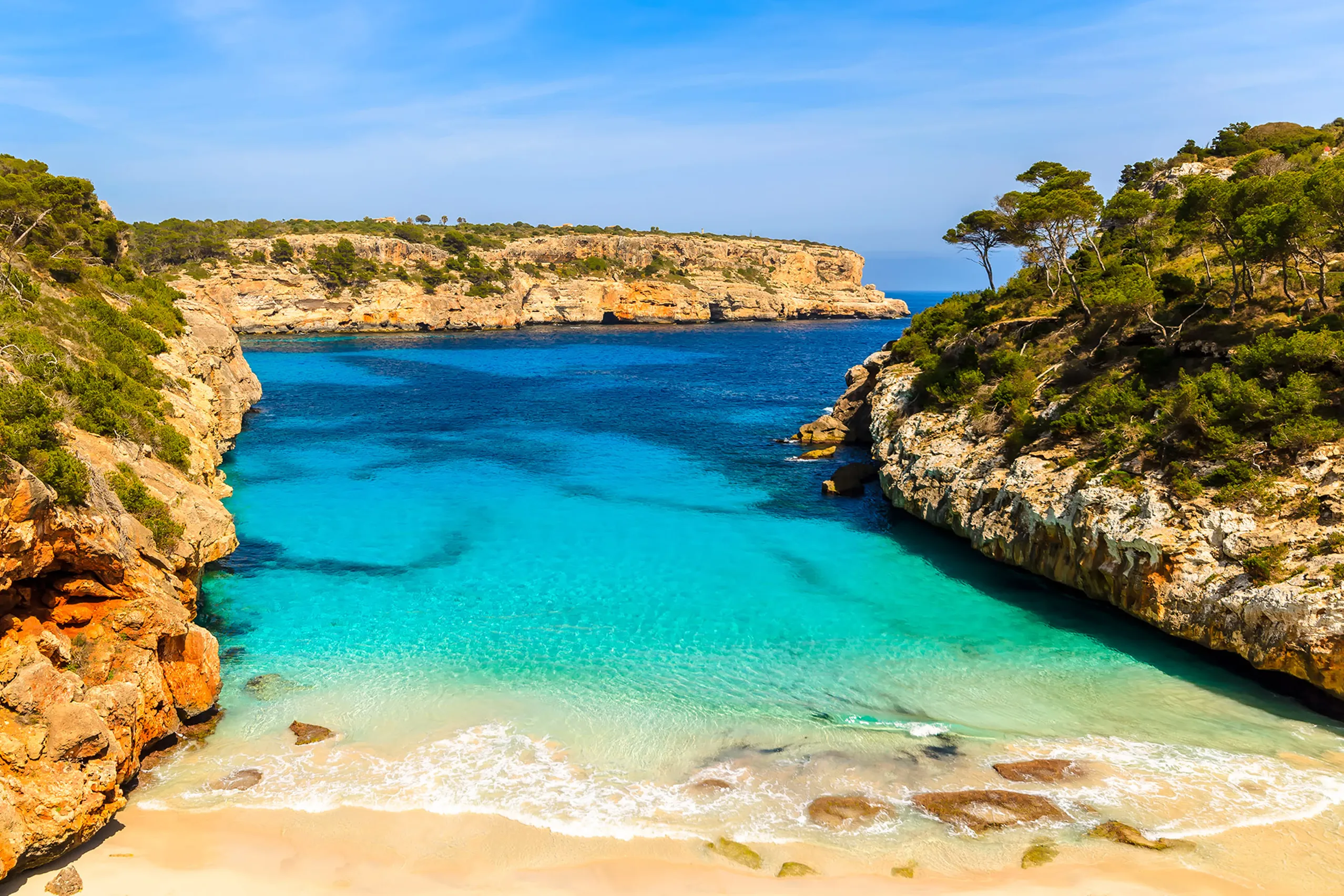 Blick von der Felskante hinunter auf den Caló des Moro: ein kleiner heller Sandstreifen zwischen steilen Felswänden, davor türkisfarbenes und tiefblaues Wasser, dahinter eine bewachsene Landzunge.
