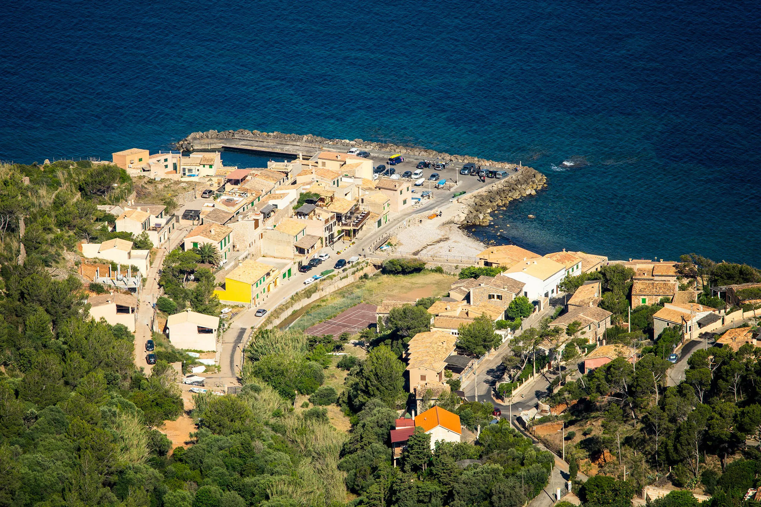 Blick von der Steilküste hinunter auf die kleine Siedlung Port de Valldemossa mit Steinhäusern, Bootsrampen und dem dunkelblauen Meer.
