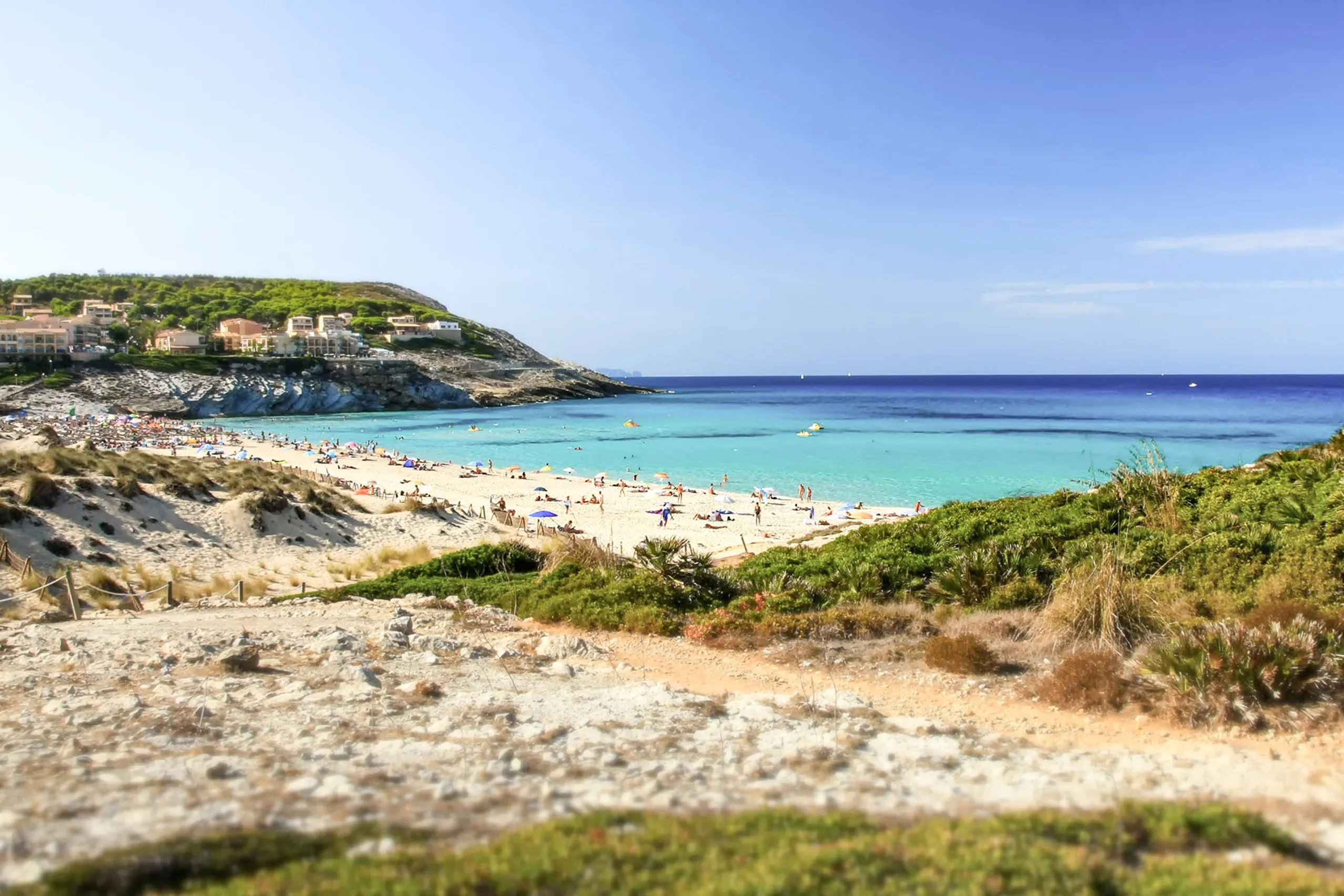 Blick von einer Anhöhe über Macchia hinweg auf die Bucht der Cala Mesquida mit hellem Sandstrand, türkisfarbenem Wasser und den weißen Häusern der Siedlung am linken Buchtrand.
