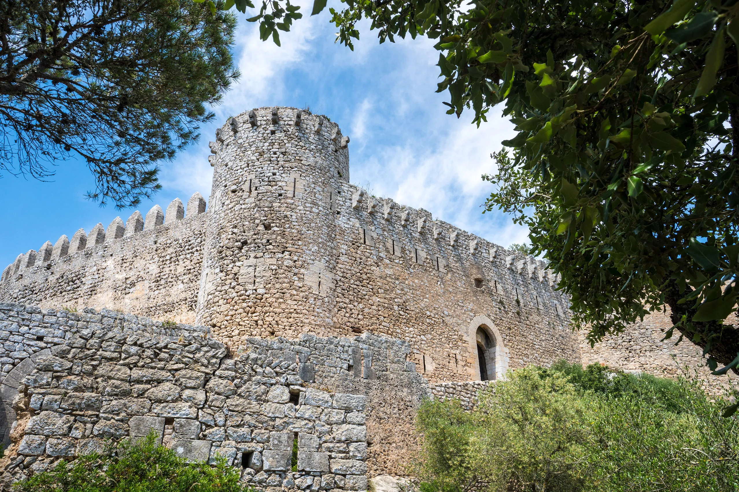 Die zinnenbewehrte Mauer des Castell de Santueri auf einem hellen Felsabbruch, dahinter blauer Himmel mit Schleierwolken.
