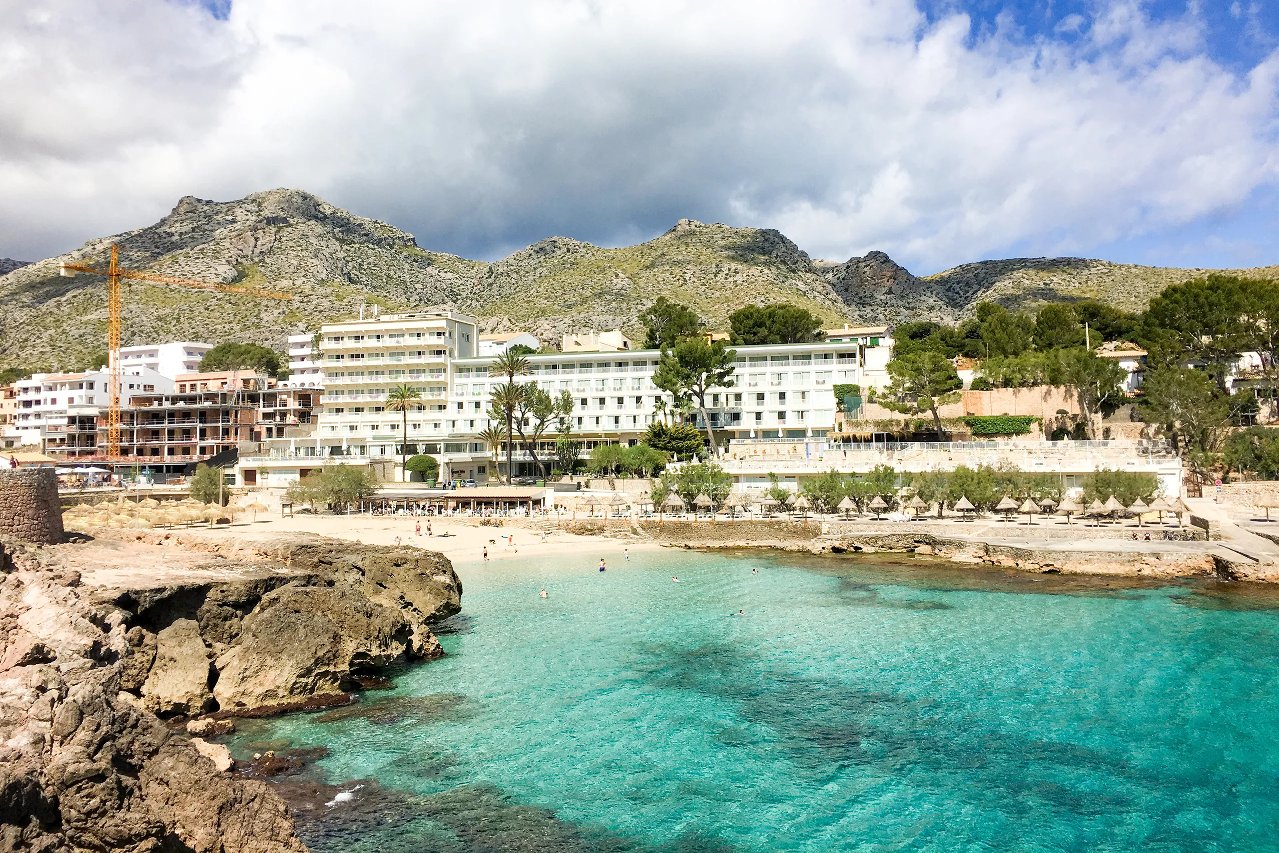 Blick von oben auf eine Bucht von Cala de Sant Vicenç mit türkisfarbenem Wasser, hellem Sandstrand mit Schirmen und den weißen Hotelbauten dahinter, im Hintergrund die kahlen Berge der Tramuntana.
