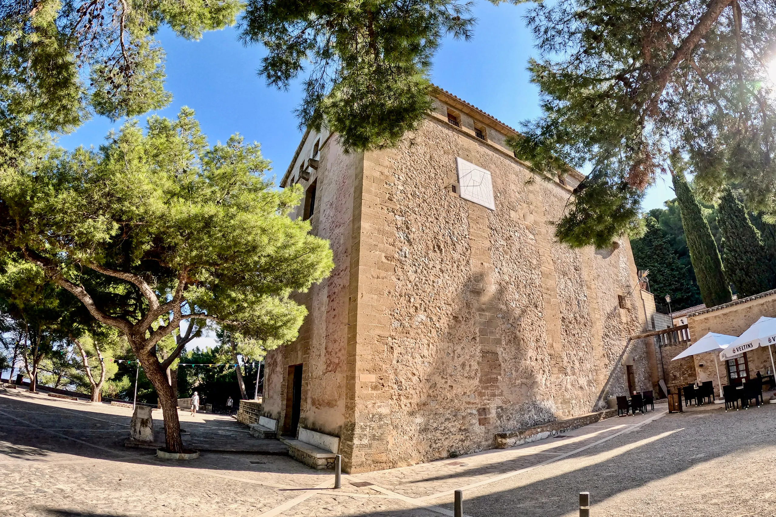 Wuchtiger Bruchsteinbau der Ermita de la Victòria mit heller Sonnenuhr an der Seitenwand, davor alte Kiefern und ein gepflasterter Vorplatz, rechts die Sonnenschirme des angrenzenden Cafés.

