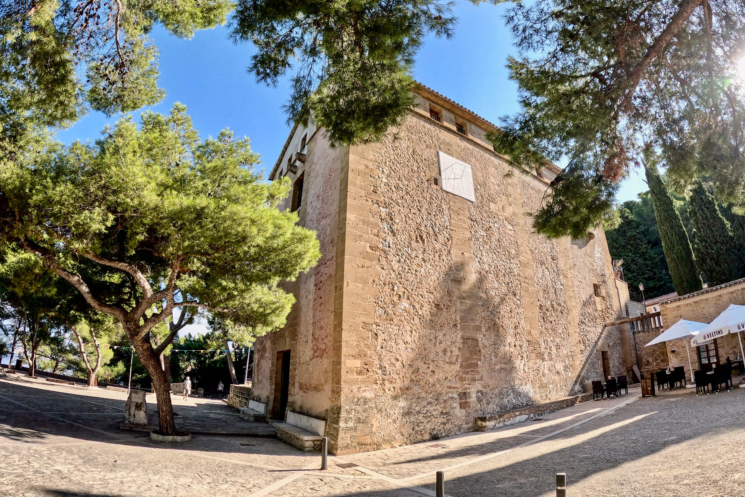 Wuchtiger Bruchsteinbau der Ermita de la Victòria mit heller Sonnenuhr an der Seitenwand, davor alte Kiefern und ein gepflasterter Vorplatz, rechts die Sonnenschirme des angrenzenden Cafés.
