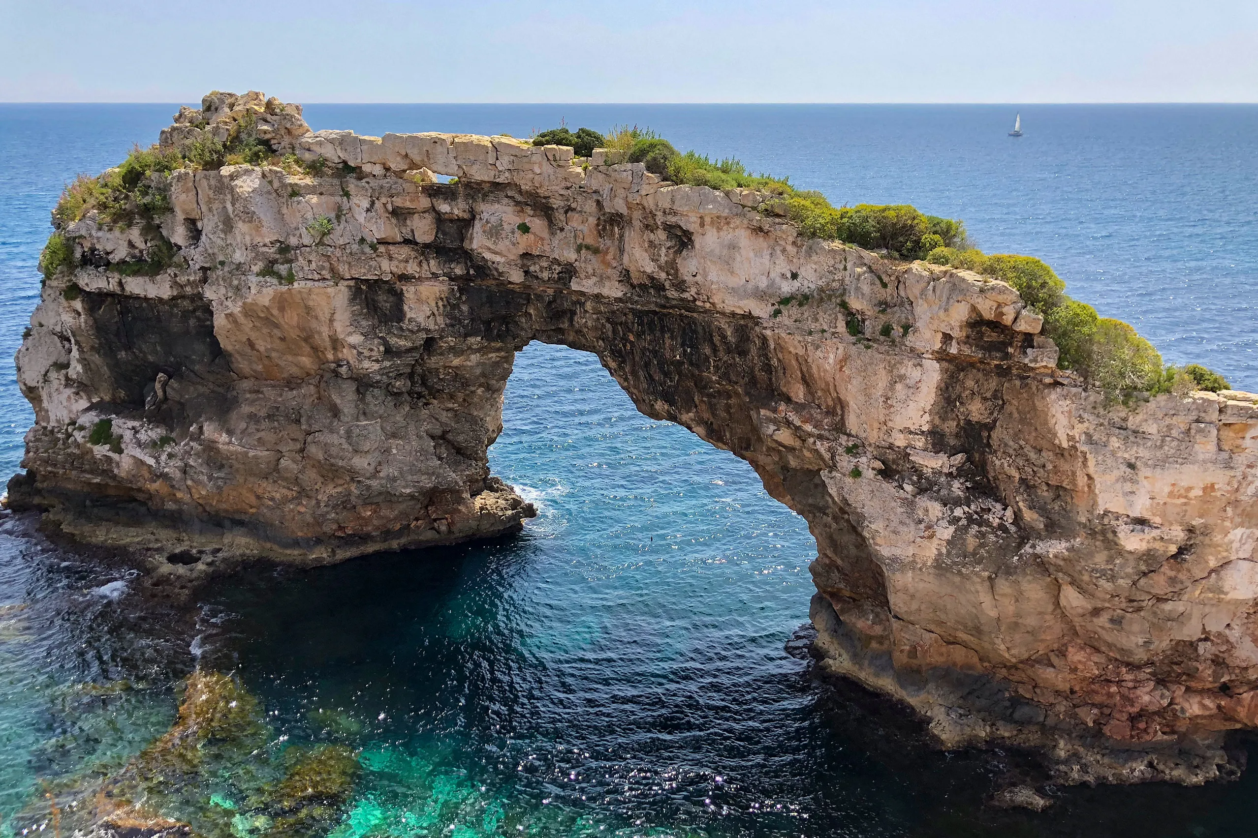 Das freistehende Felsentor Es Pontàs im türkisfarbenen Meer vor der Küste bei Santanyí, mit bewachsener Kuppe über dem Bogen und der Steilküste dahinter.
