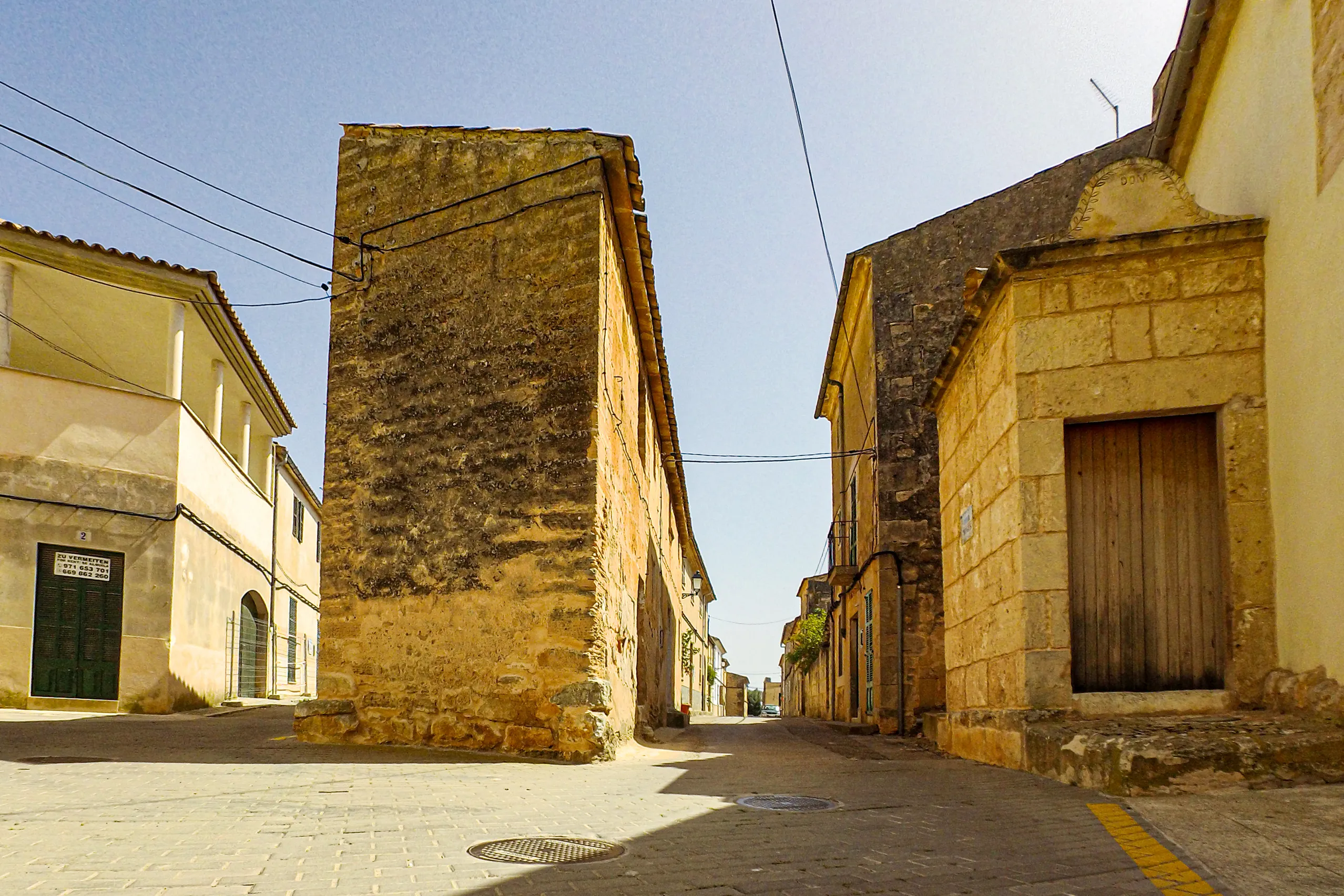Hohe fensterlose Giebelwand aus Bruchstein an einer Gassengabelung in Ses Salines, rechts ein Haus mit steinernem Portal.
