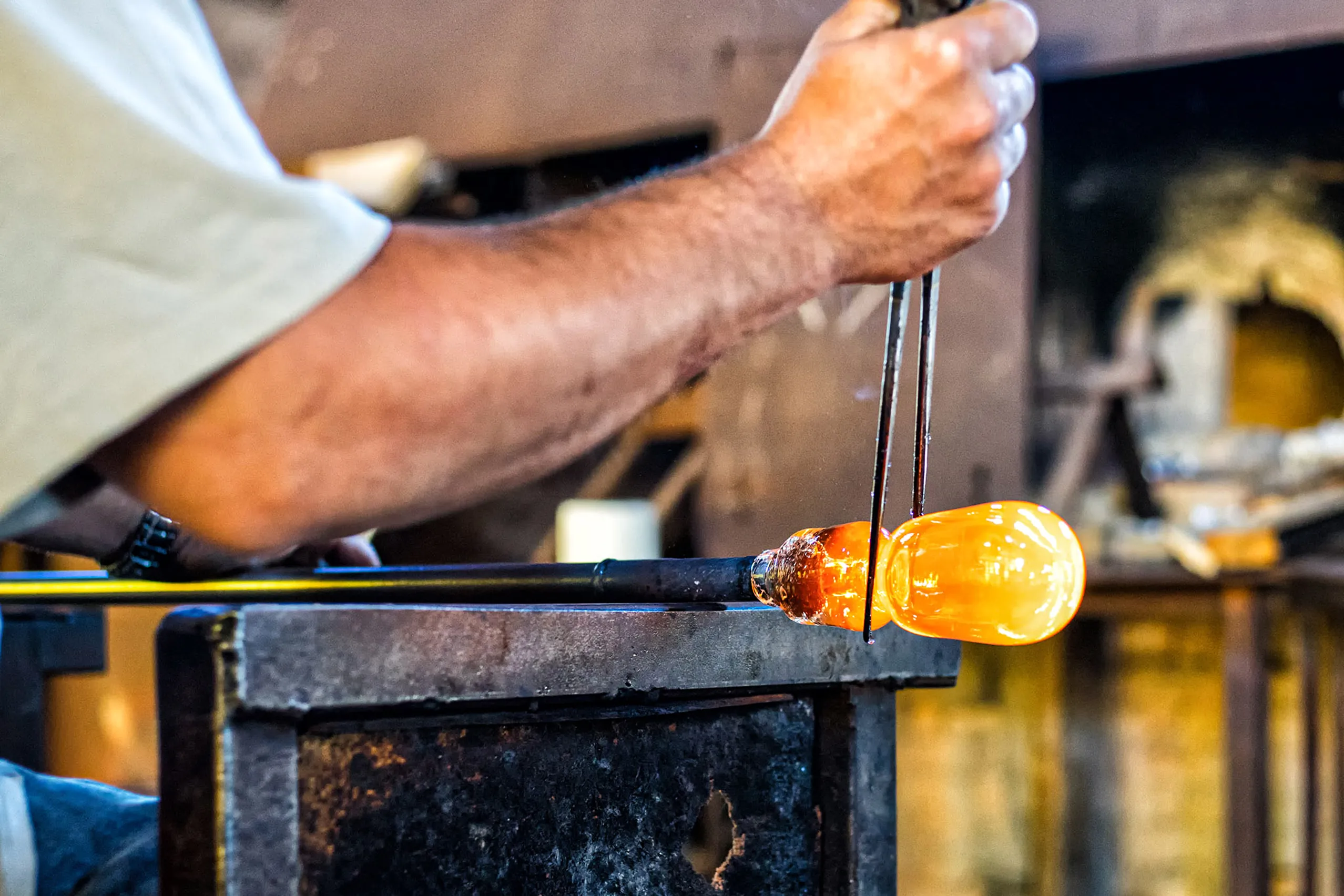 Hände eines Glasbläsers, die eine Pfeife mit glühendem, orange leuchtendem Glas über einer Werkbank drehen.
