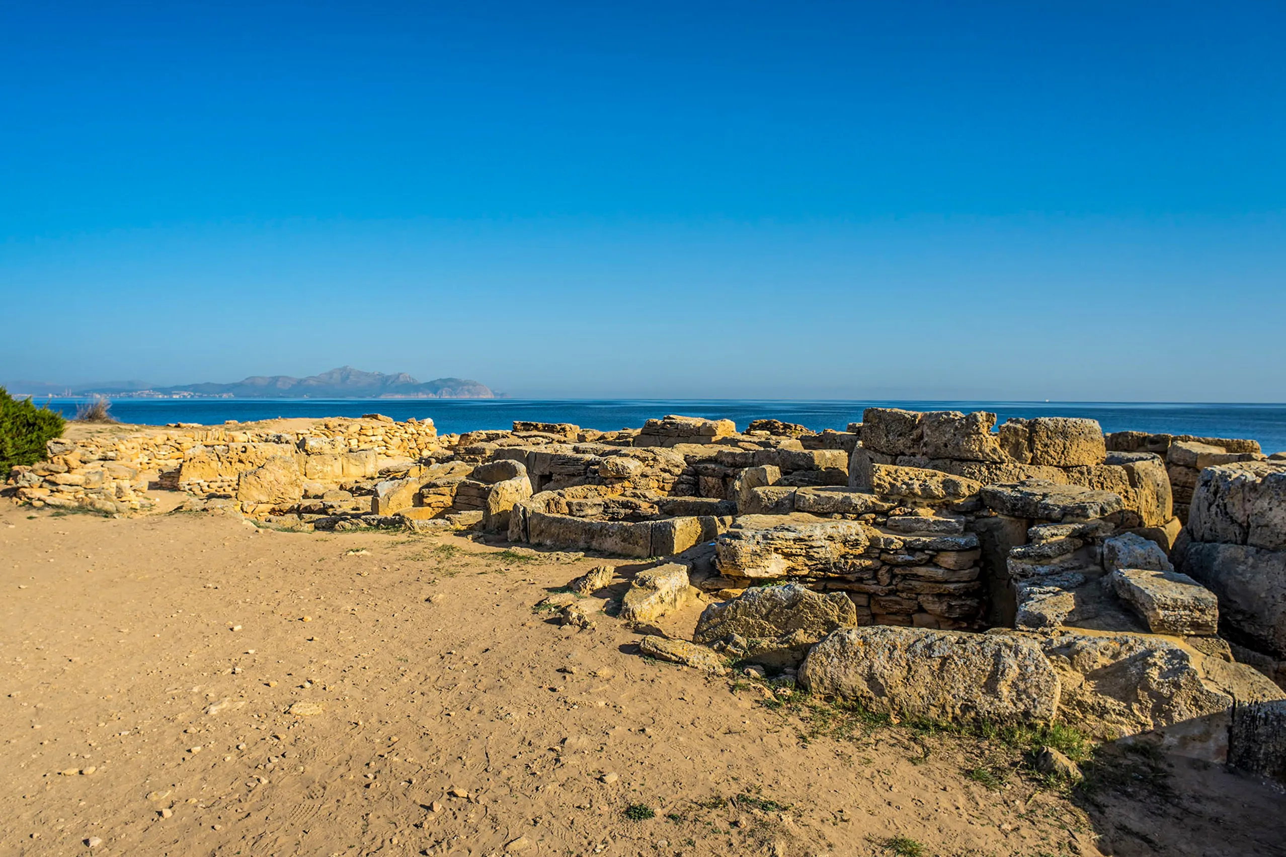 Das talayotische Gräberfeld von Son Real unmittelbar an der Küste, helle Steinkammern im Sand, dahinter das türkisfarbene Meer und die Halbinsel am Horizont.
