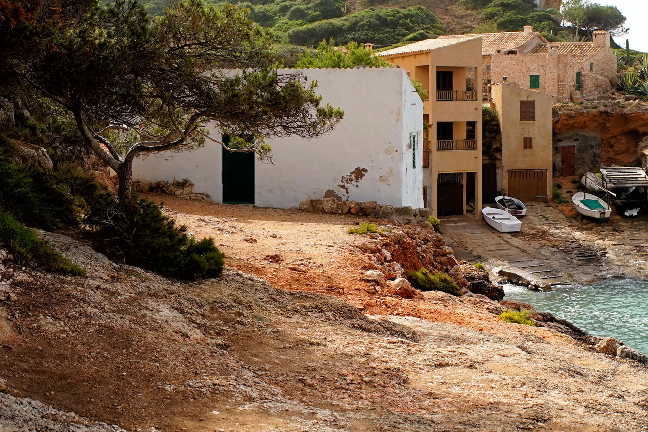 Alte Fischer- und Bootshäuser aus hellem Sandstein über der Felsbucht Cala s'Almunia, davor türkisfarbenes Wasser und blanker Fels.
