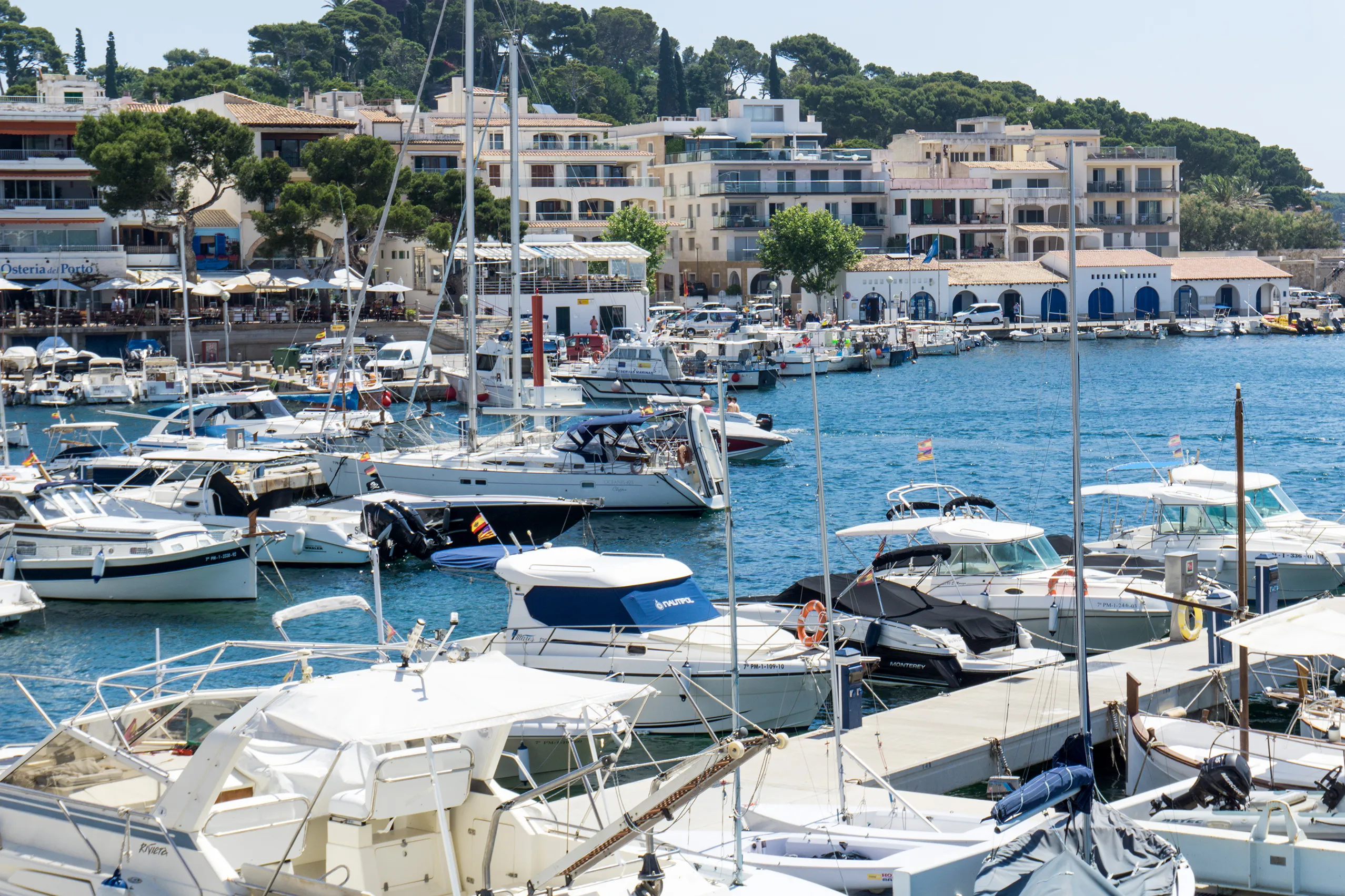 Der Hafen von Cala Ratjada mit dicht liegenden weißen Motorbooten, dahinter die Häuser- und Lokalzeile am Kai und bewaldete Hügel.
