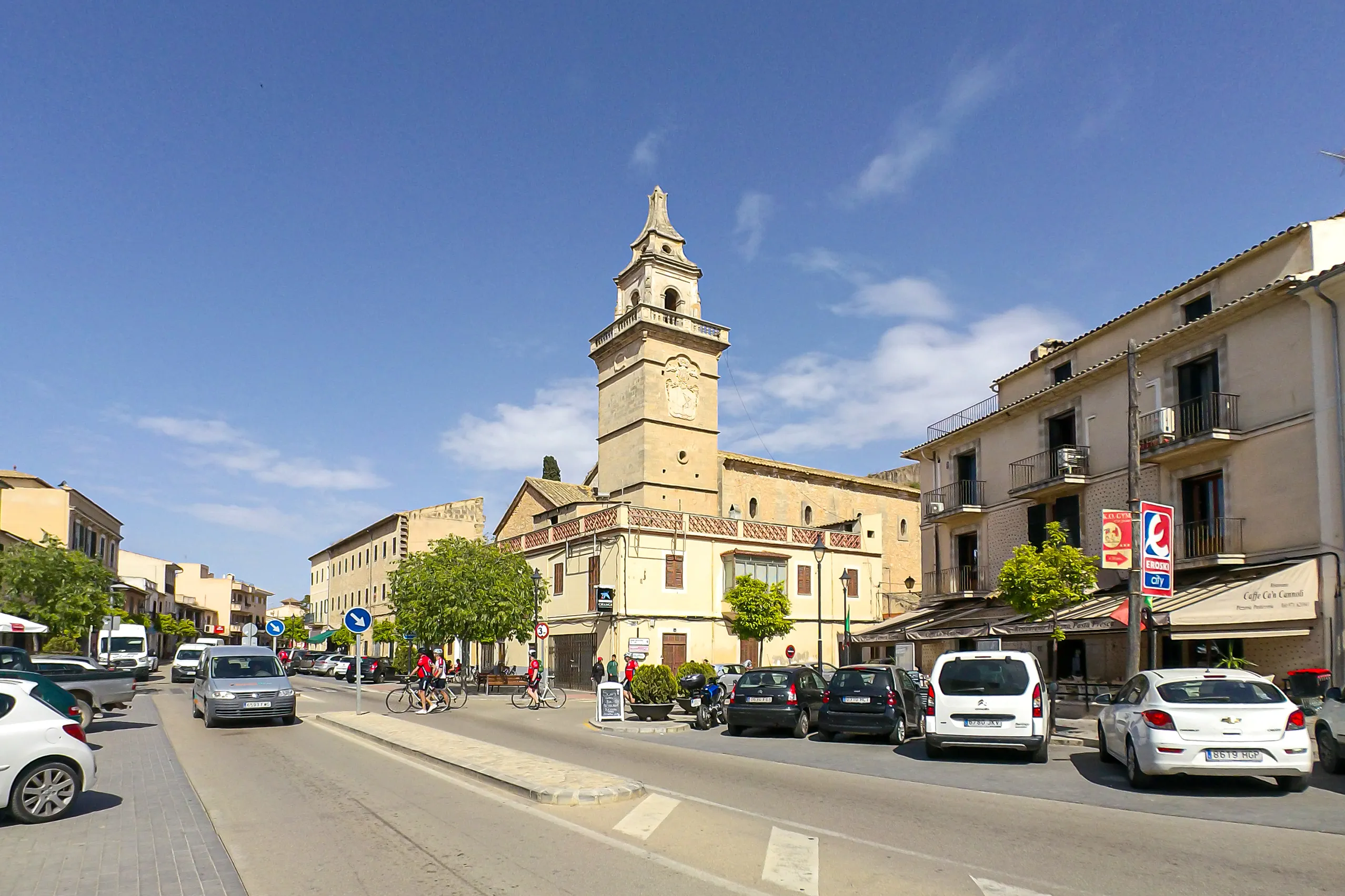 Die Durchgangsstraße von Santa Maria del Camí mit parkenden Autos, Häuserzeilen und dem markanten Uhrturm der Kirche im Hintergrund.
