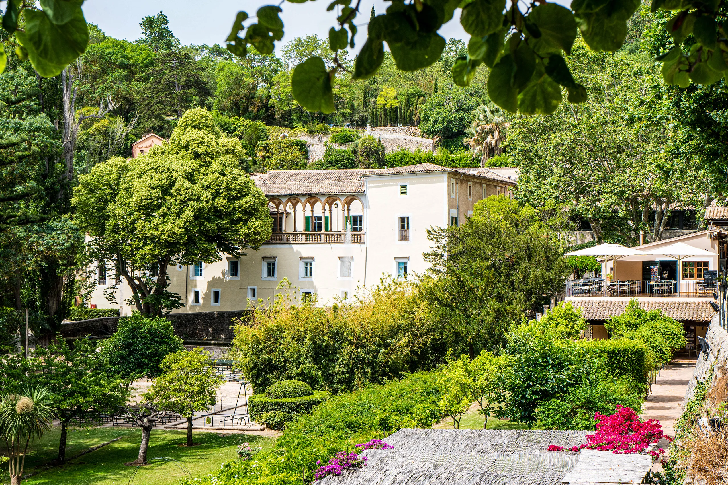 Das Herrenhaus von La Granja mit heller Fassade und Loggia, eingebettet in dichte Bäume und Gärten am Hang der Serra de Tramuntana.
