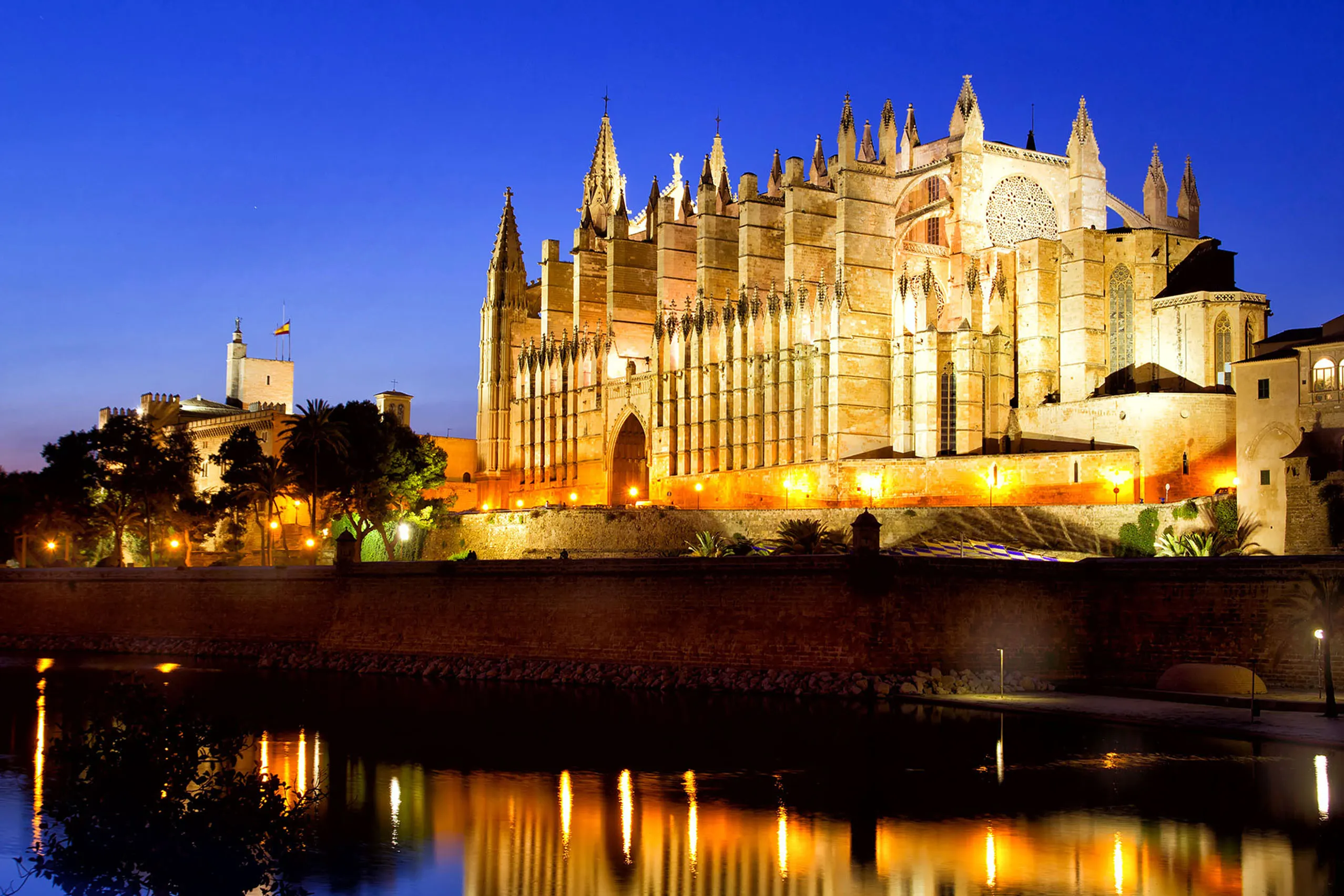 Die angestrahlte Kathedrale von Palma zur blauen Stunde, davor der Palau de l'Almudaina, im Vordergrund die Spiegelung im Wasserbecken des Parc de la Mar.
