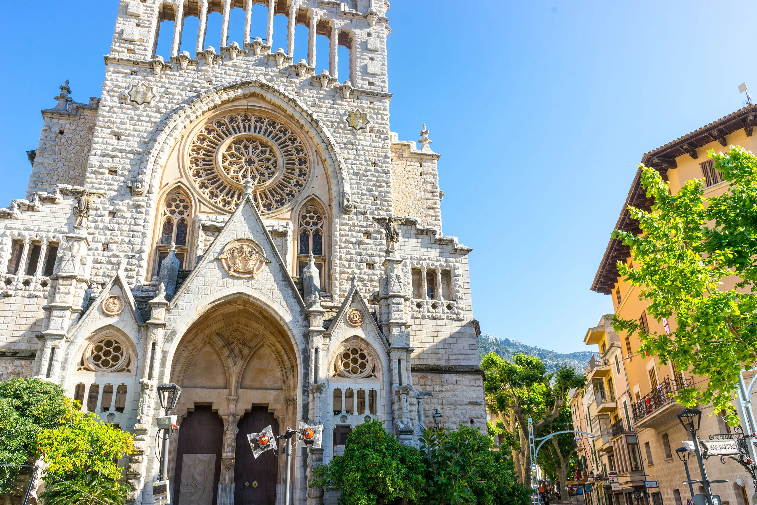 Der Hauptplatz von Sóller mit der Kirche Sant Bartomeu, dem Rathaus mit Fahnen und Platanen, davor Passanten und ein geparktes Auto.
