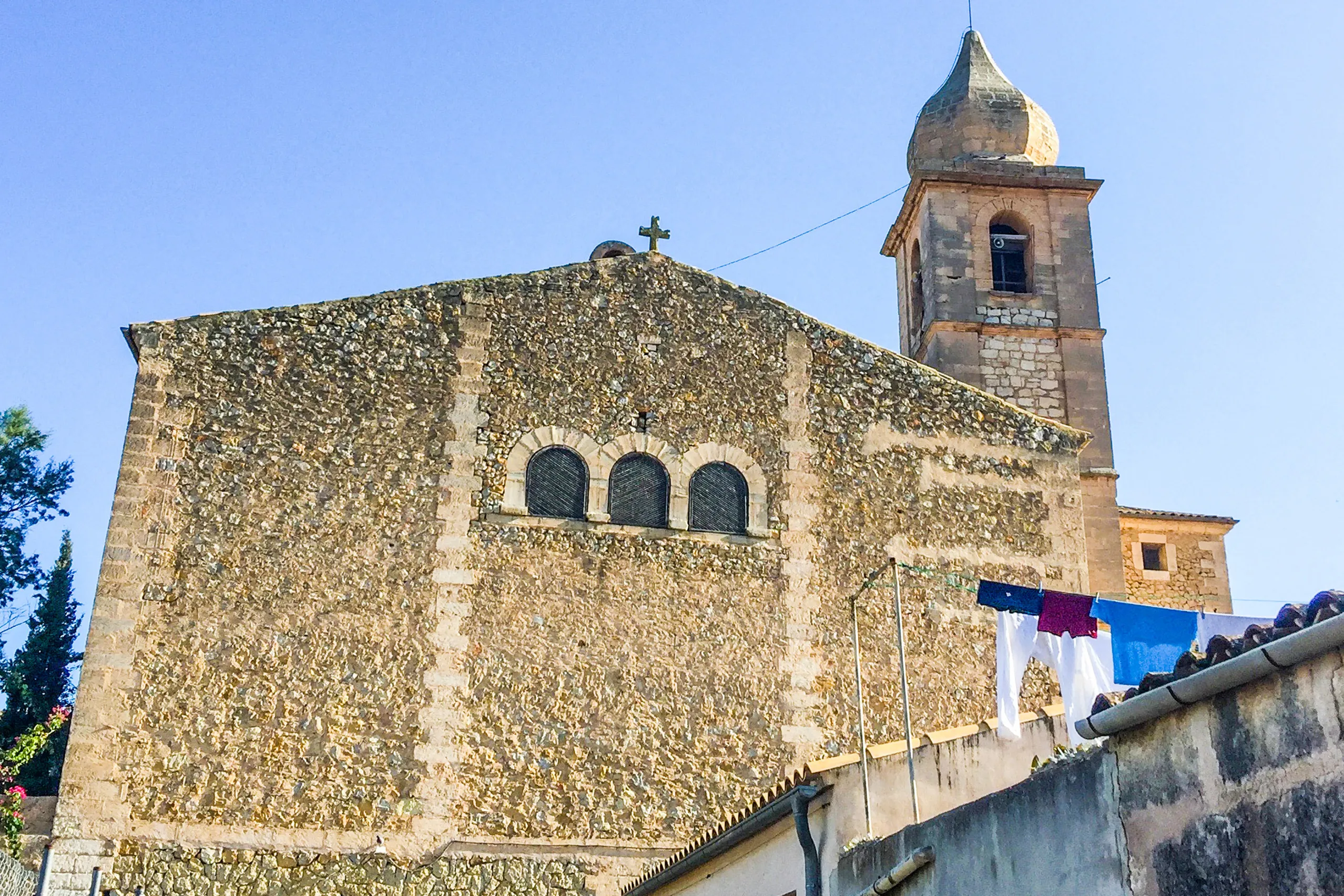 Die wuchtige Pfarrkirche von Maria de la Salut aus unregelmäßigem Feldstein mit hohem Glockenturm, davor niedrige Wohnhäuser.
