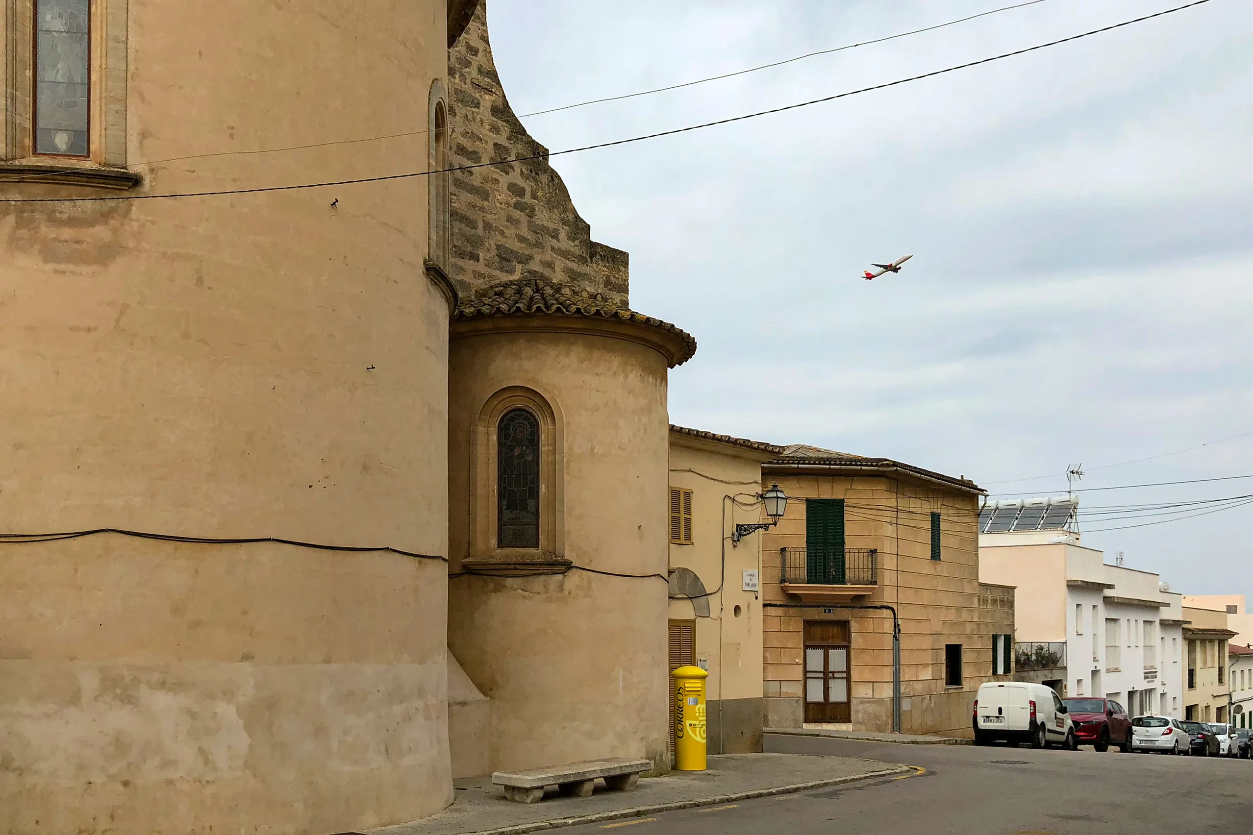 Die Apsis der Kirche von Sant Jordi im Vordergrund, dahinter Dorfhäuser, ein gelber Briefkasten und ein startendes Flugzeug am Himmel.

