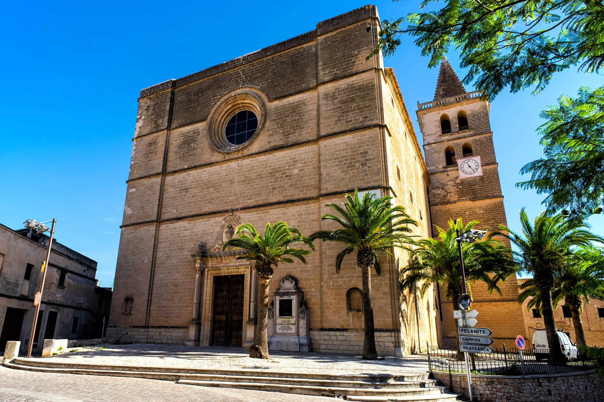 Die mächtige Kirche von Porreres aus hellem Stein mit großem Rundfenster, barockem Portal und hohem Glockenturm, davor Palmen und ein gepflasterter Vorplatz.
