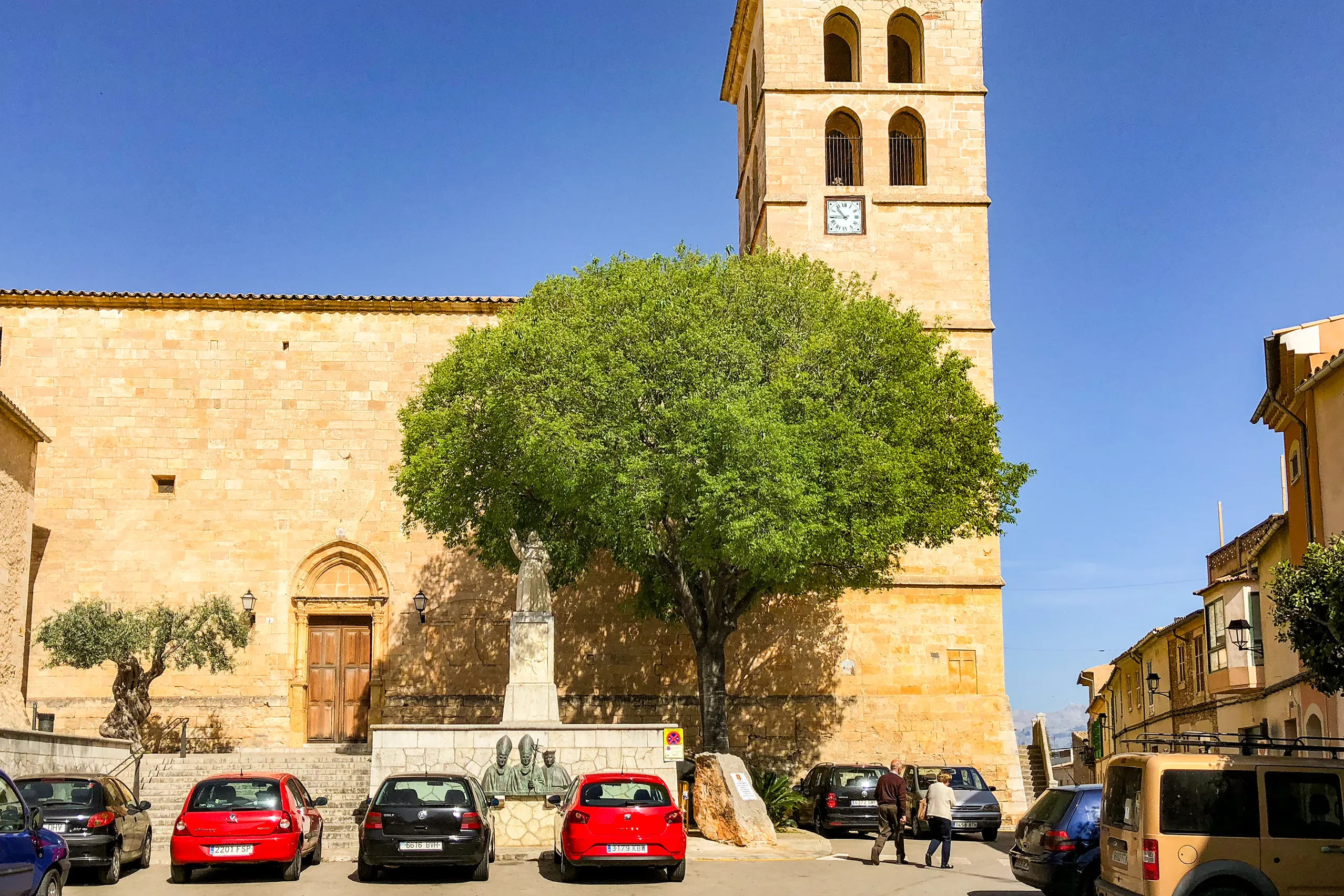 Die Pfarrkirche von Sencelles aus hellem Sandstein mit hohem Glockenturm, davor ein Platz mit Baum, Denkmal und parkenden Autos.
