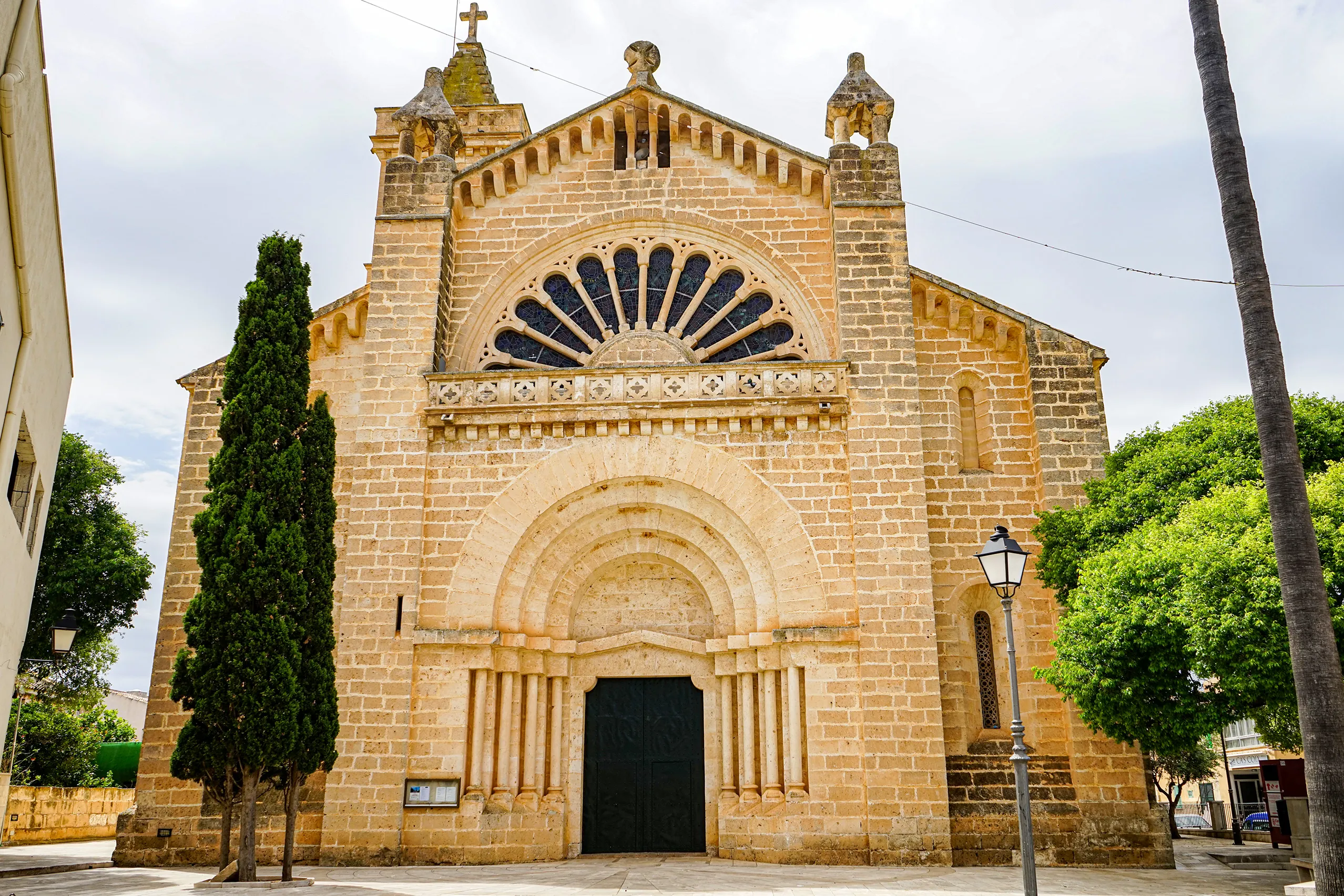 Die Kirche Sant Miquel in Son Carrió aus hellem Sandstein mit Rundbogenportal, halbrunder Fächerrosette und zwei kleinen Türmchen, davor eine Zypresse.
