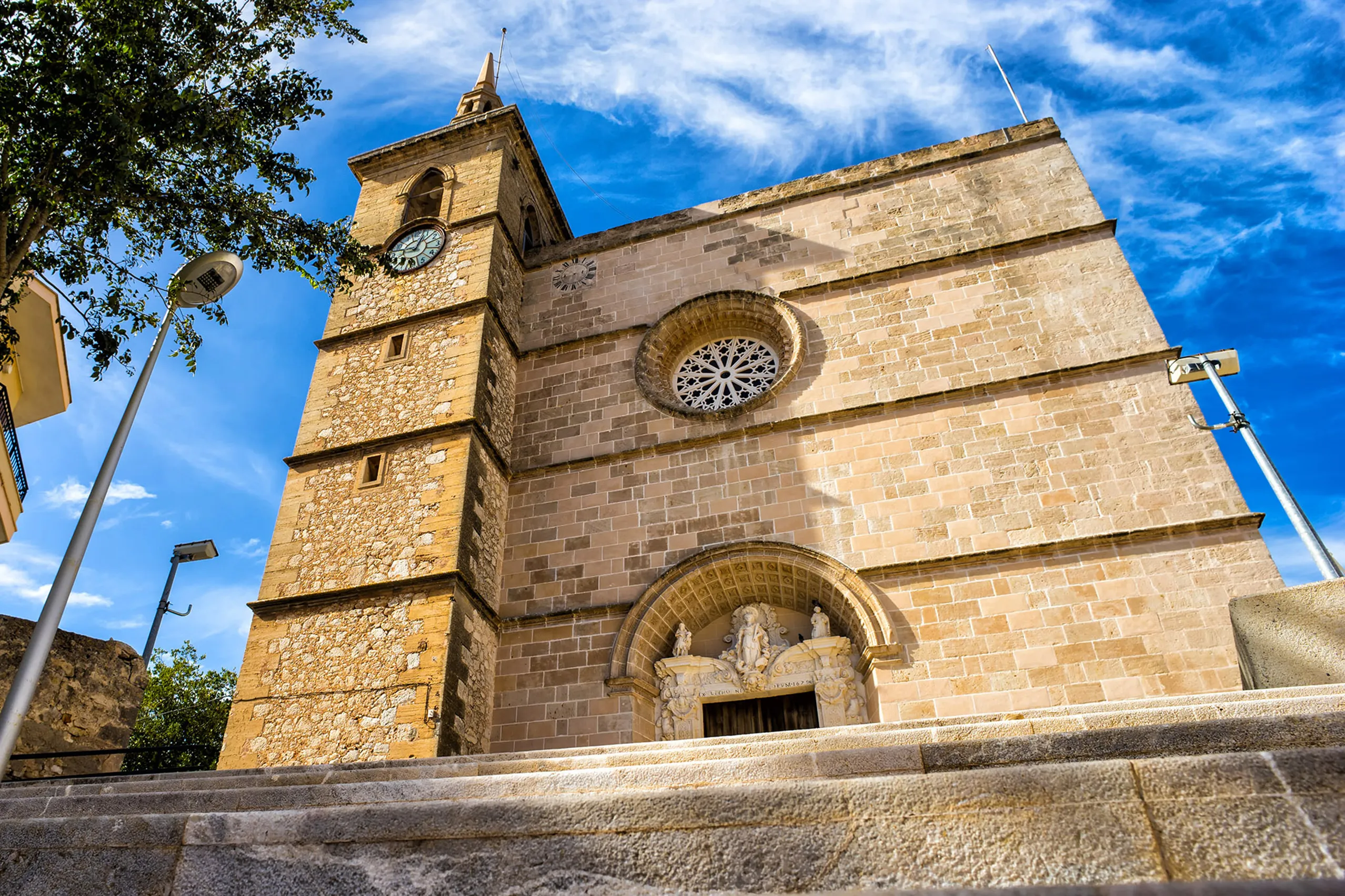 Die Pfarrkirche von Santa Margalida aus hellem Naturstein mit Rundfenster und wuchtigem Glockenturm, davor Bäume und ein blauer Himmel.
