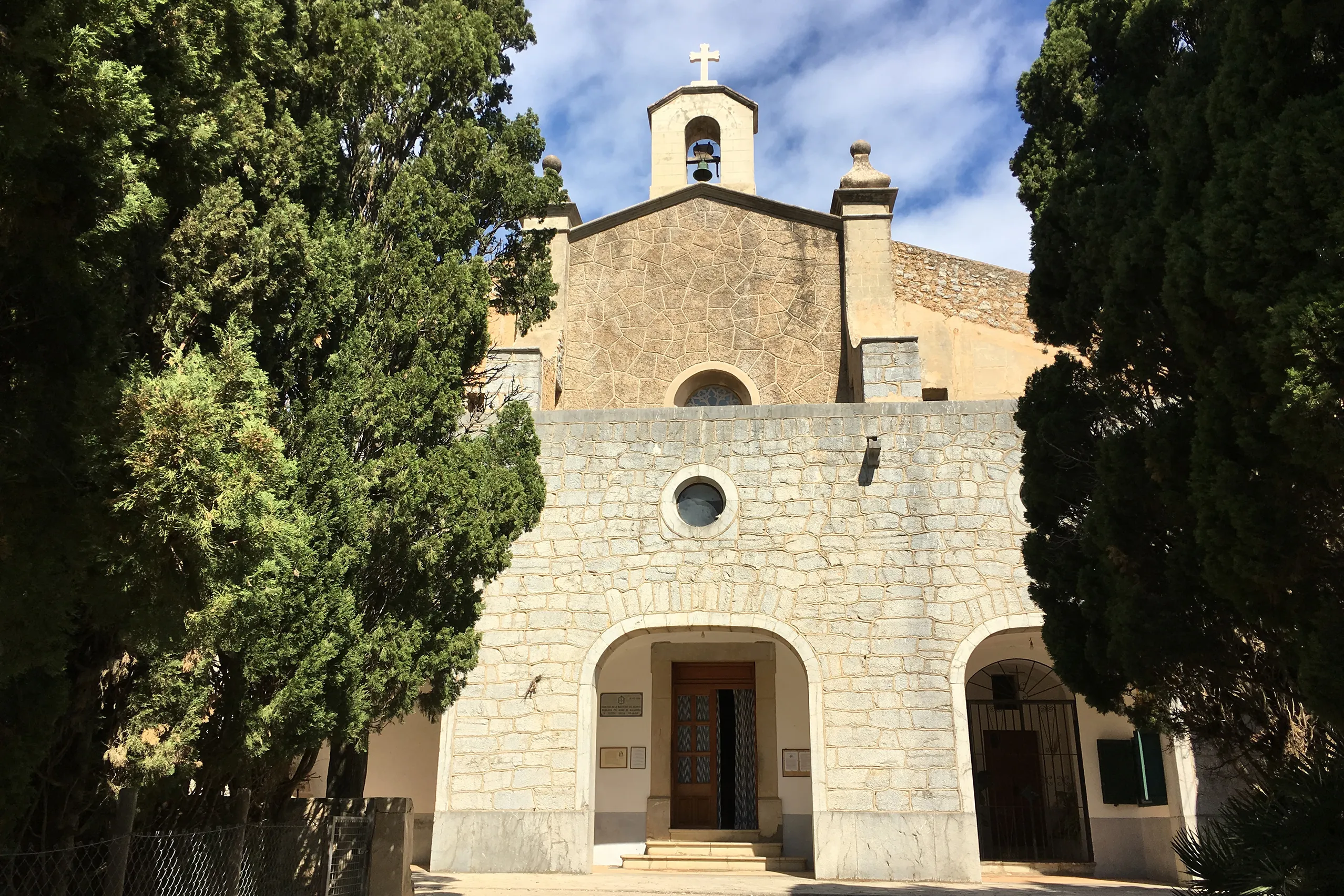 Die Kirche der Ermita de Betlem aus hellem Bruchstein mit Glockengiebel, Rundbogenportal und einer runden Fensteröffnung, eingerahmt von hohen dunklen Zypressen.
