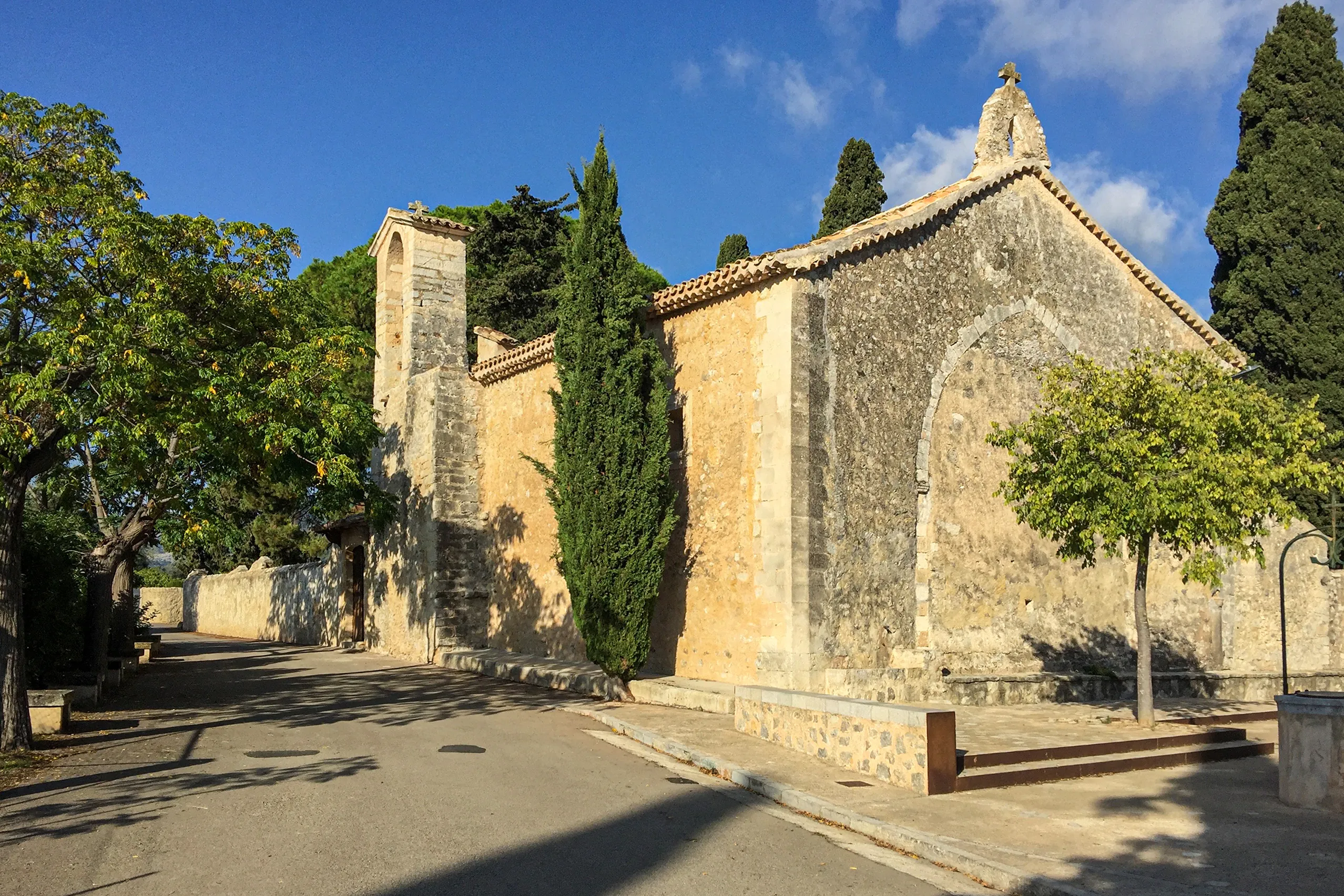 Die alte Kirche Sant Miquel bei Campanet aus hellem Naturstein mit Glockengiebel, flankiert von Zypressen, unter blauem Himmel.
