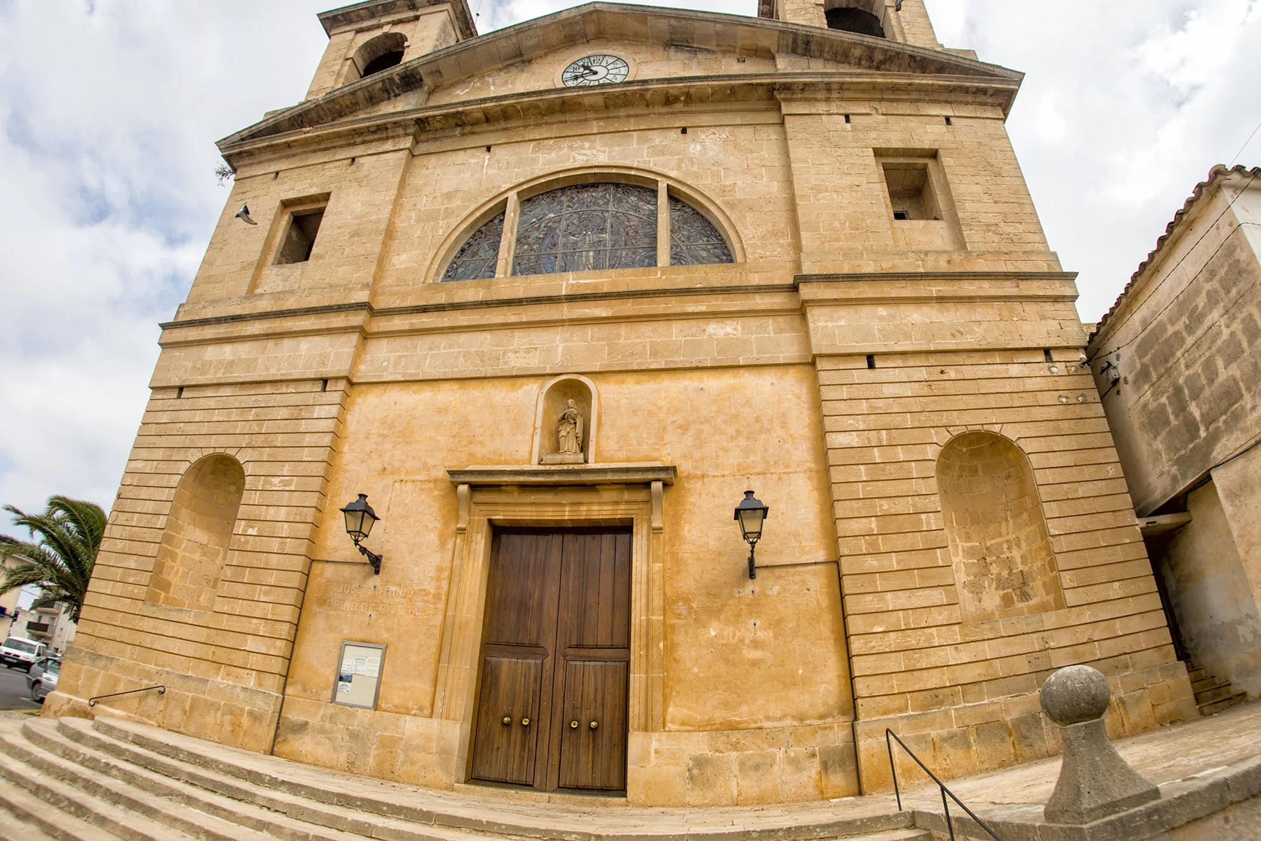 Die Fassade der Pfarrkirche von S'Alqueria Blanca aus gelbem Sandstein mit hohem Bogenfenster, Uhr, Heiligennische und hölzernem Portal, davor eine breite Treppe.
