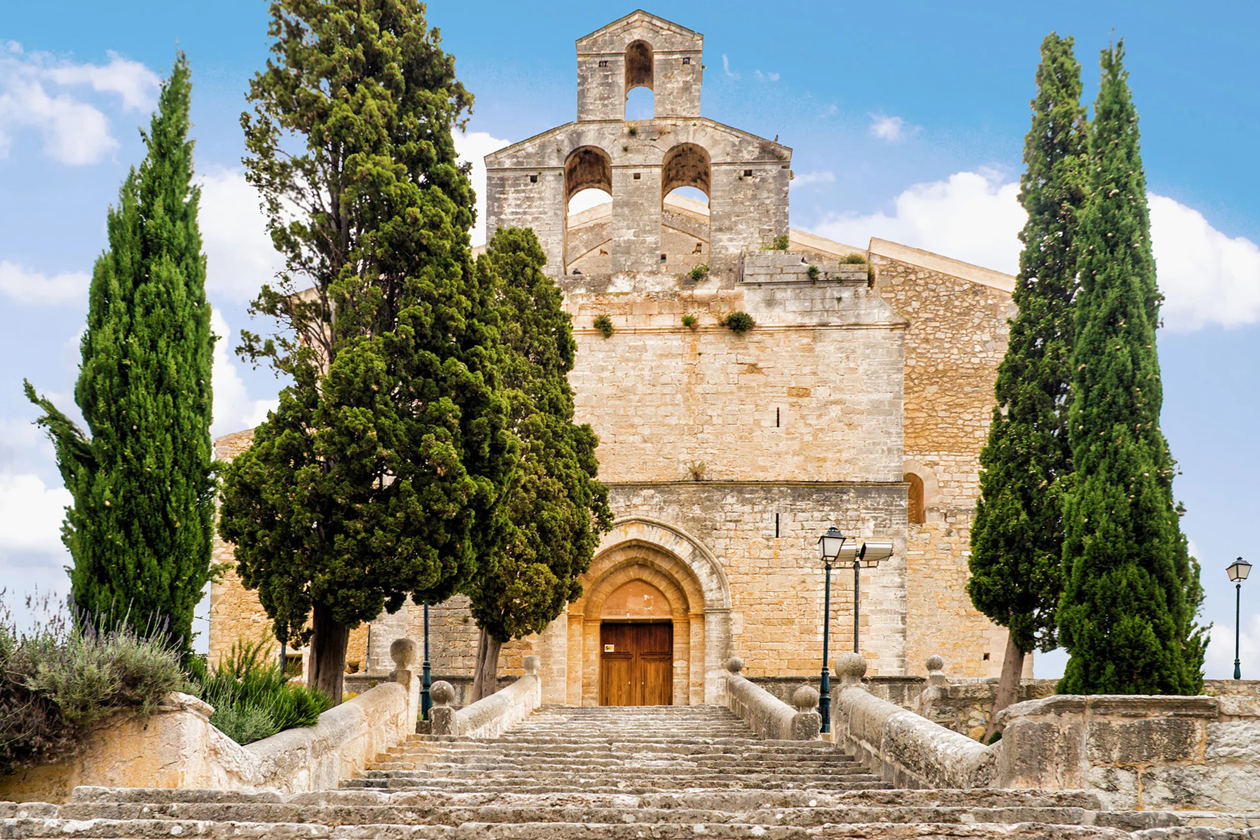 Die Kirche Sant Llorenç in Selva aus hellem Naturstein mit Glockengiebel und gotischem Portal, davor eine breite steinerne Freitreppe zwischen hohen Zypressen.

