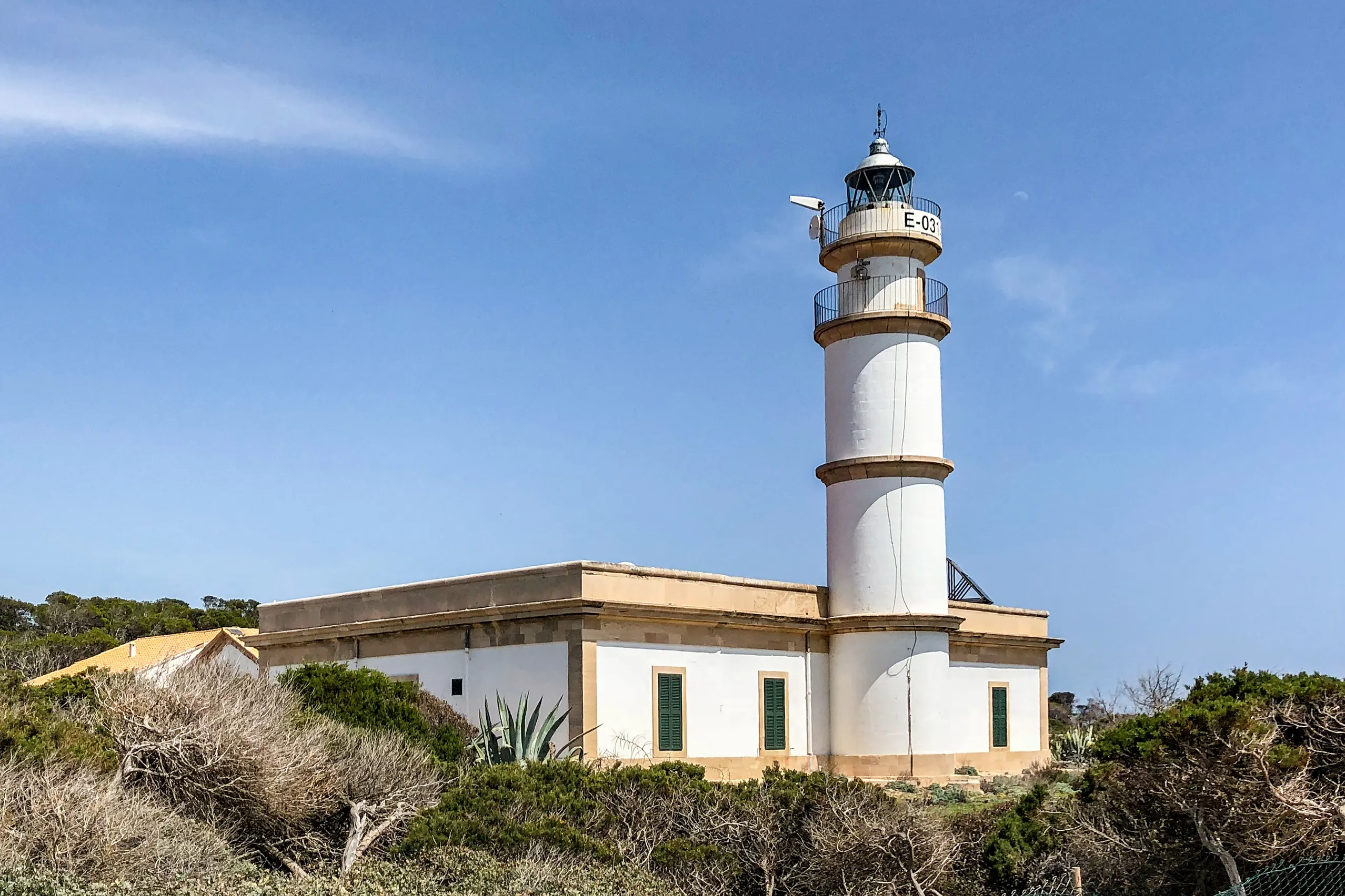 Der weiße Leuchtturm am Cap de ses Salines mit dem niedrigen Wärterhaus, davor zerklüftete helle Felsplatten und das Meer.
