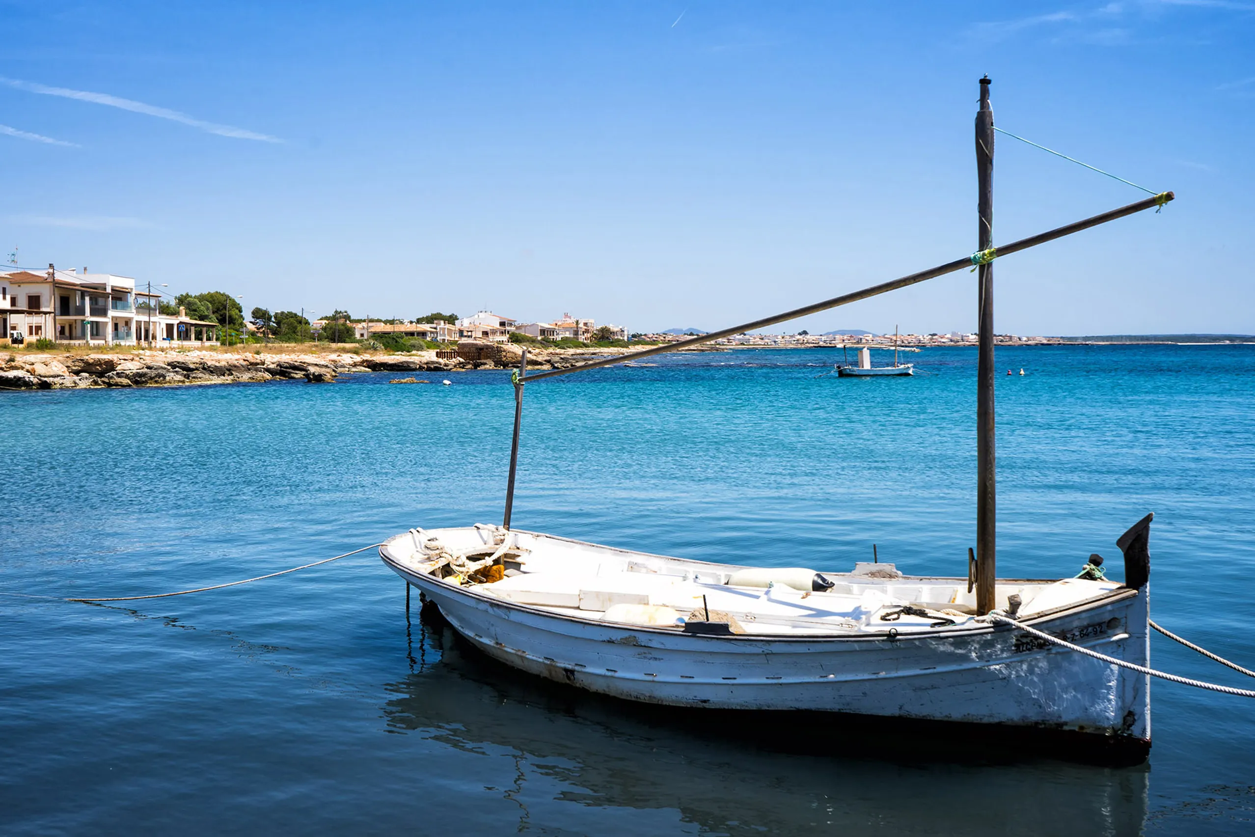 Weißes hölzernes Boot mit langem Mast im klaren blauen Wasser vor S'Estanyol de Migjorn, am Ufer flache Häuser und Kiefern.
