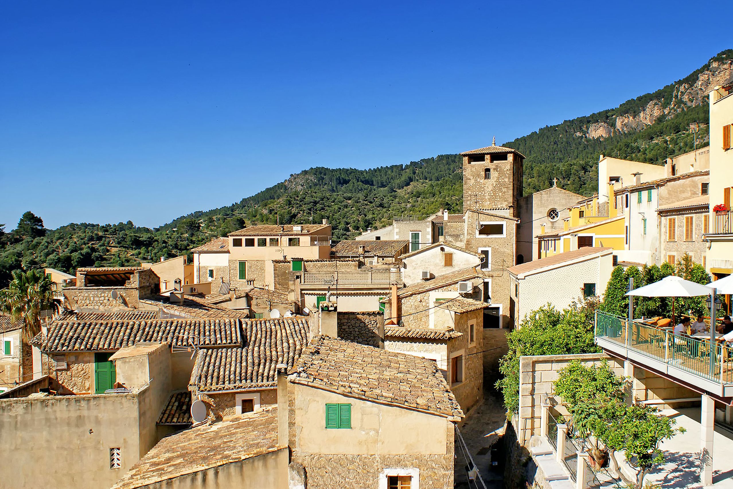 Blick über die Ziegeldächer von Estellencs am Steilhang, dazwischen der wehrhafte Kirchturm, im Hintergrund die bewaldeten Hänge der Tramuntana.
