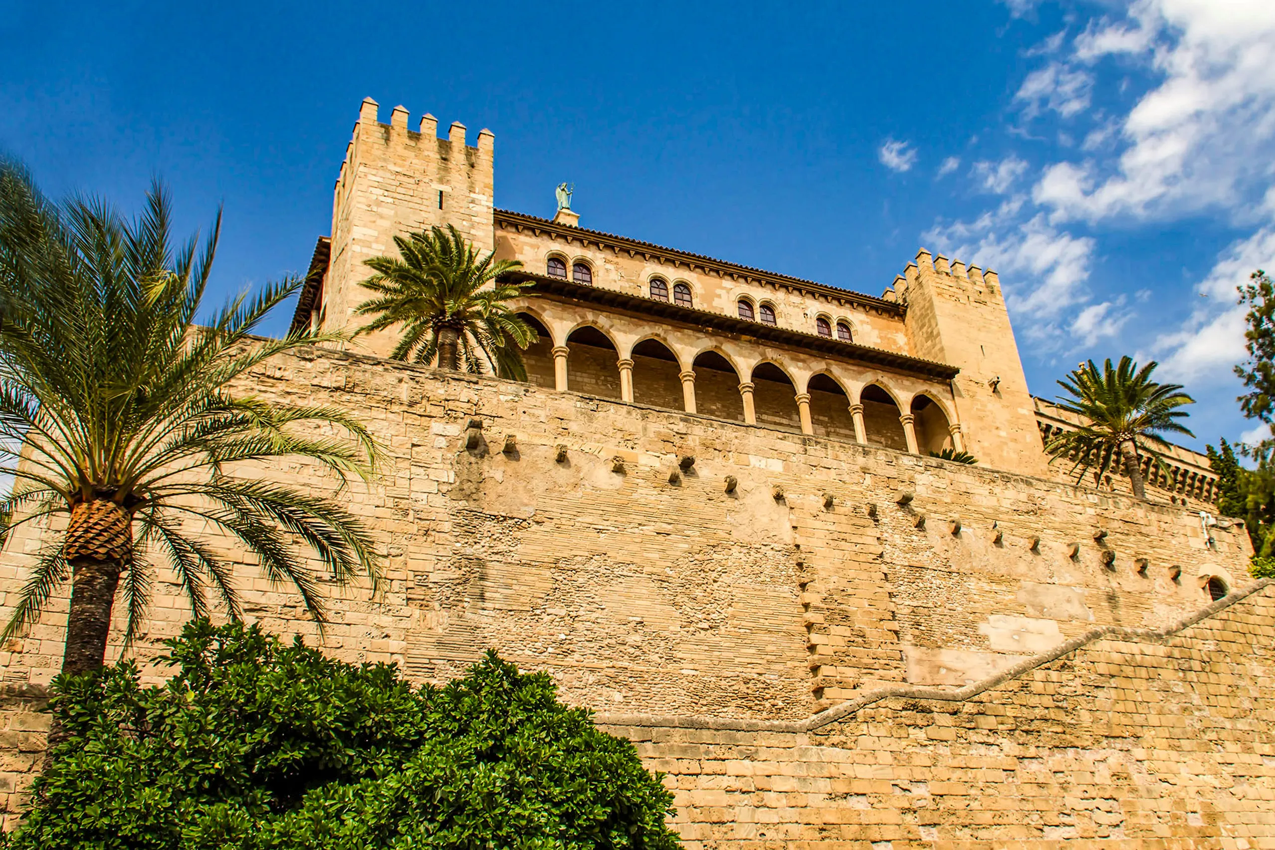 Der Palau de l'Almudaina in Palma mit Zinnenturm, Arkadenreihe und Sandsteinmauer, davor Palmen und Zypressen unter blauem Himmel.
