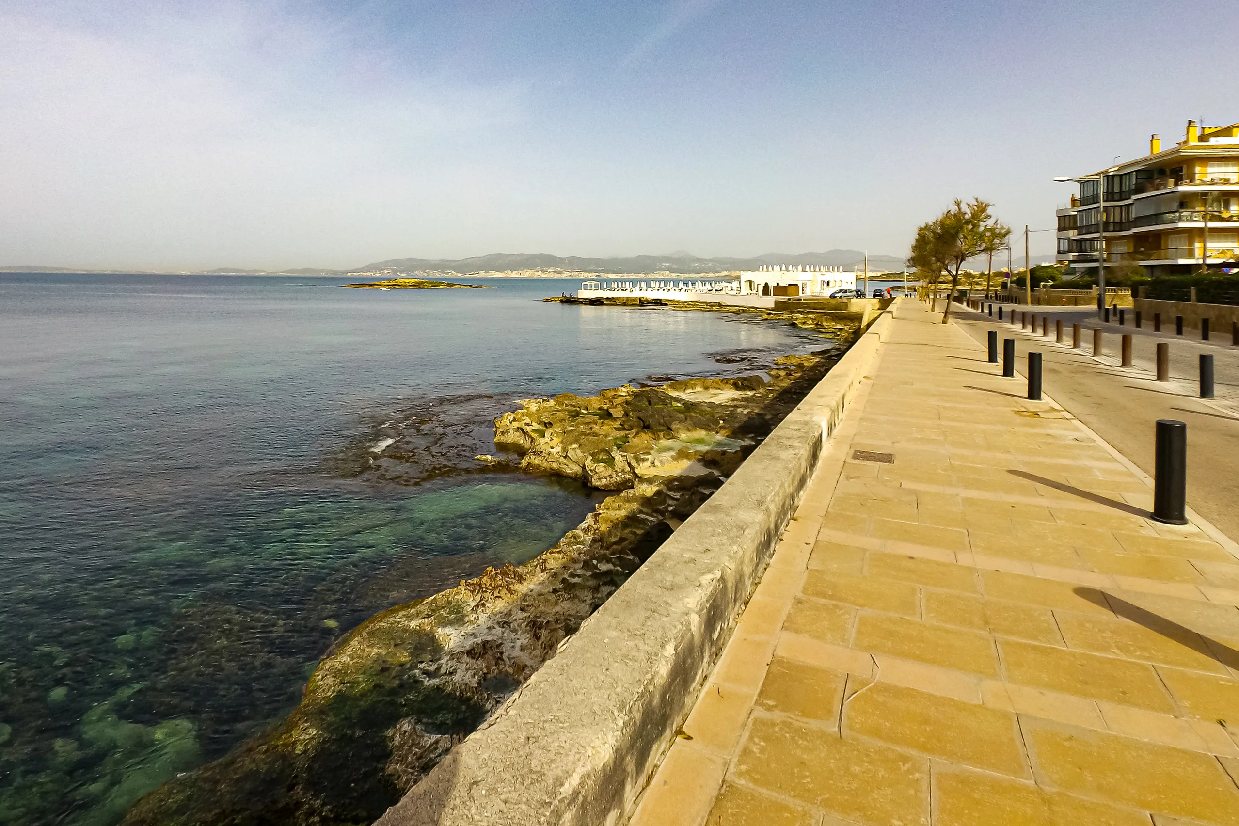 Gepflasterte Uferpromenade in Can Pastilla entlang der flachen Felsküste mit klarem Wasser, dahinter die Bucht von Palma.
