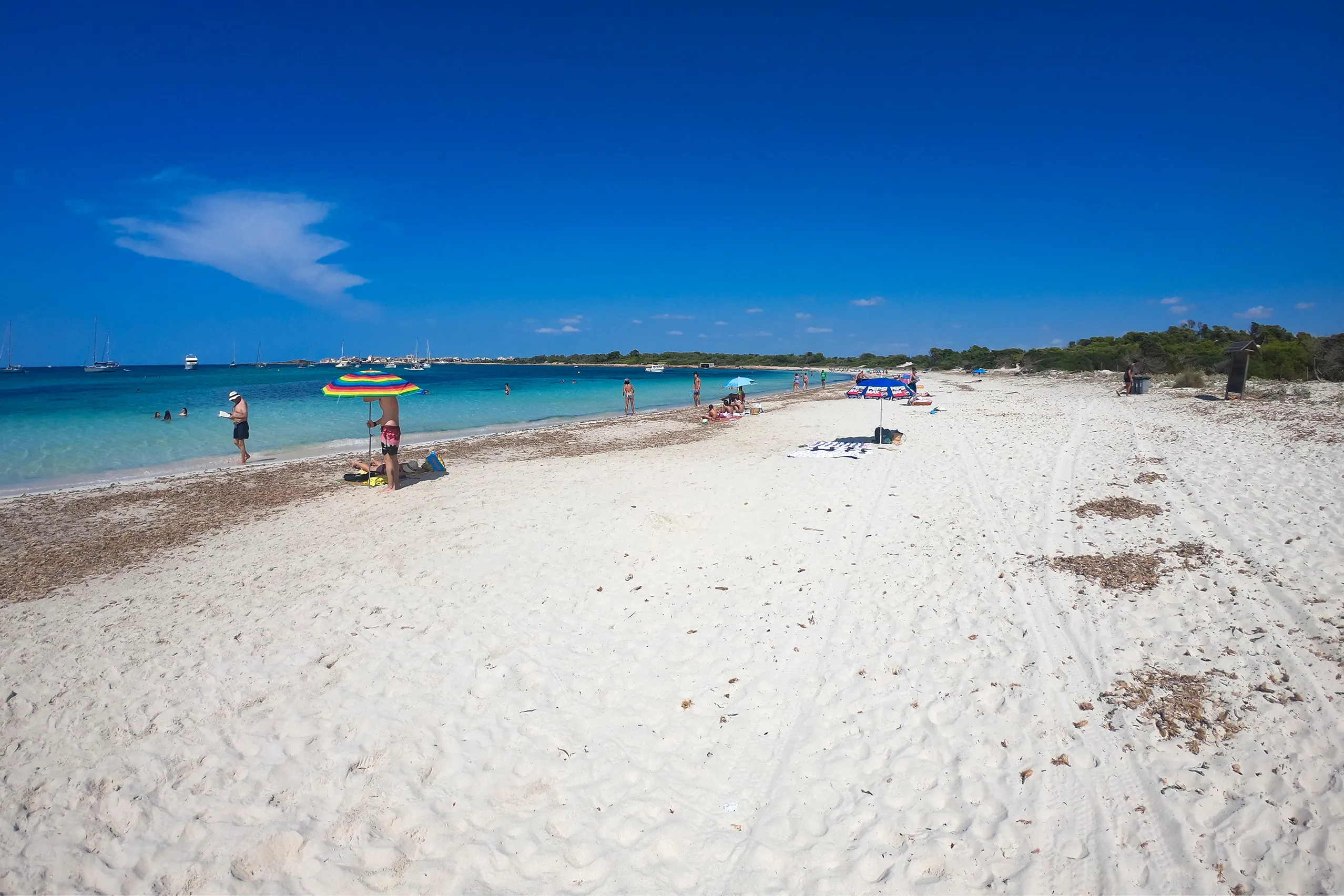 Sehr klares, flach abfallendes Wasser an einem schmalen weißen Sandstreifen der Platja des Carbó, im Hintergrund Kiefern und ankernde Boote.
