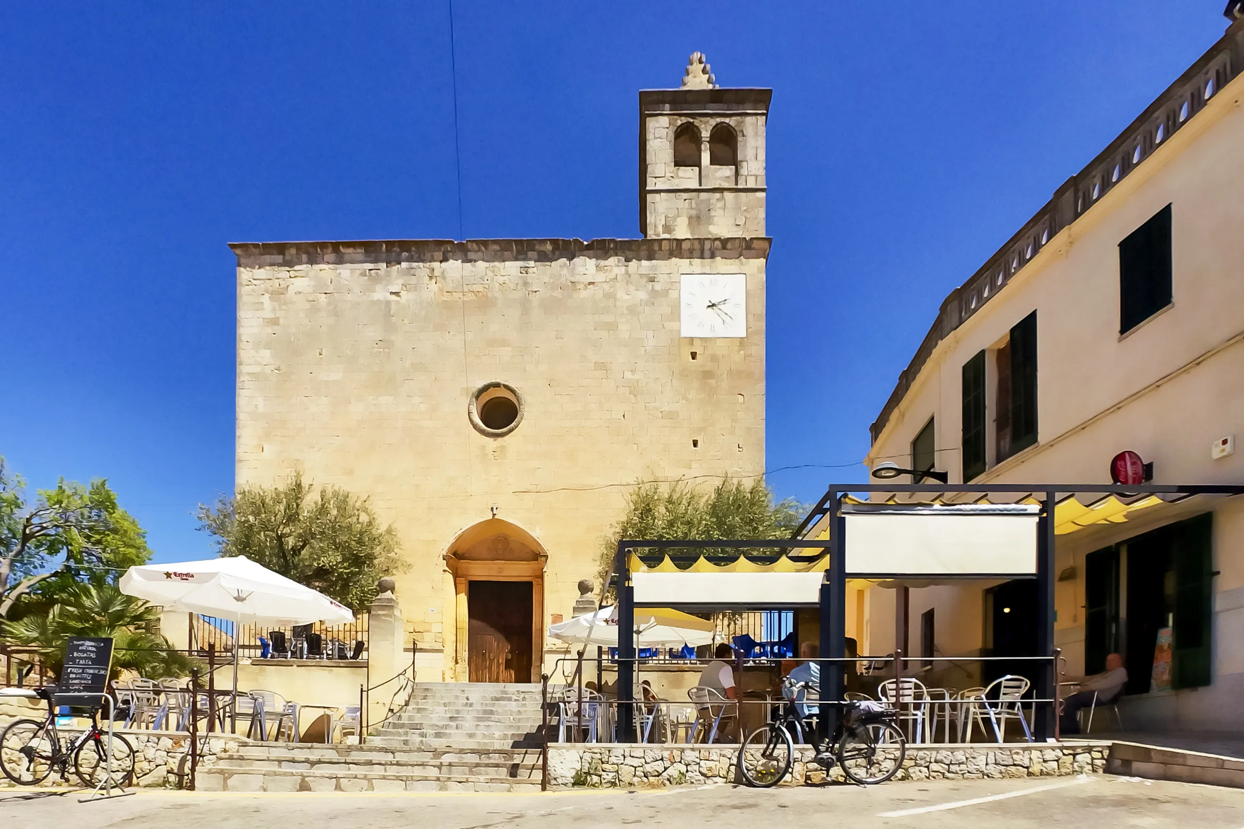 Die Pfarrkirche Sant Pere in Búger aus hellem Naturstein mit schlichtem Glockenturm, davor ein Platz mit Bäumen, Café-Terrasse und geparkten Fahrrädern.
