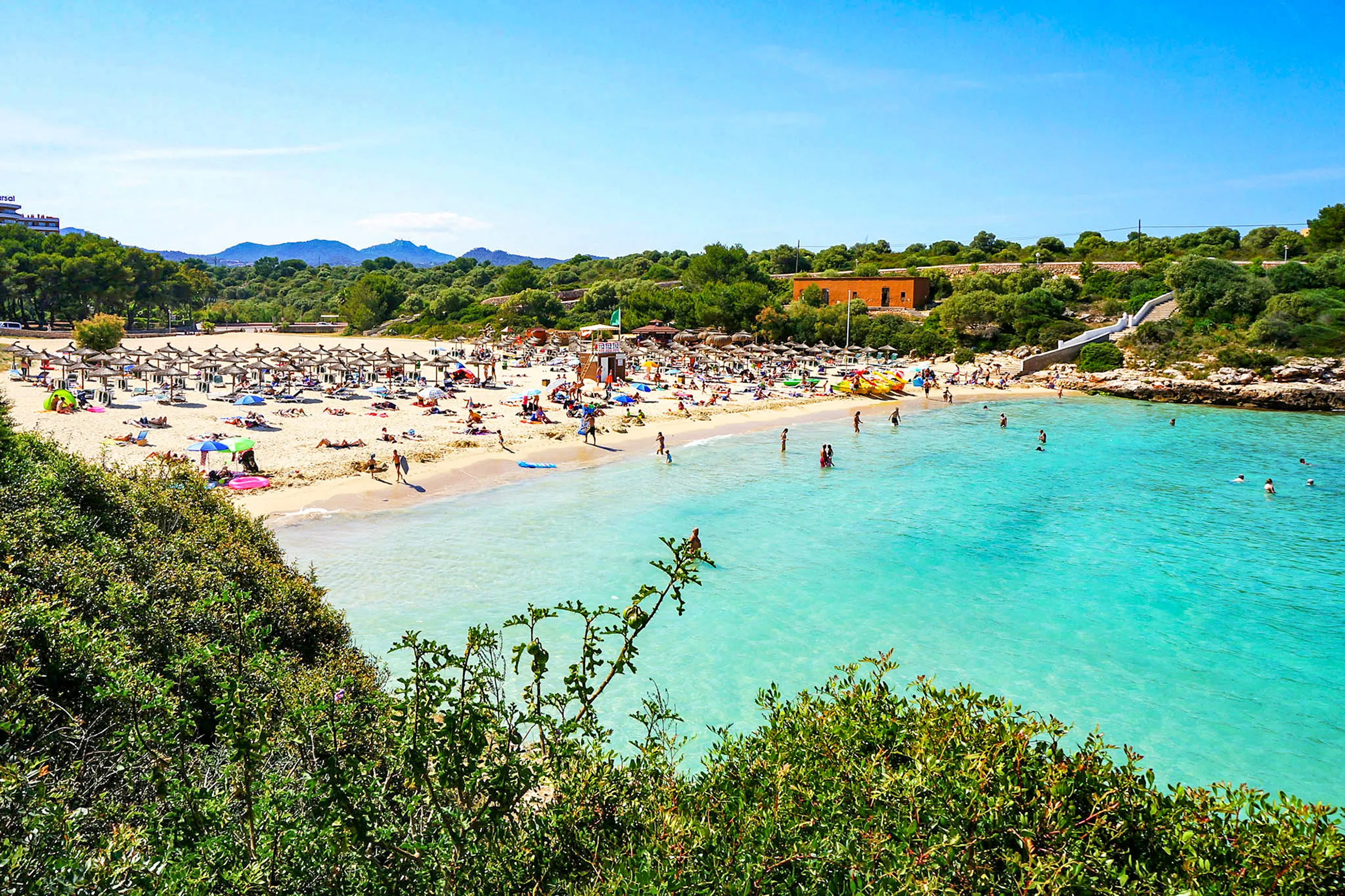 Dicht belegter goldgelber Sandstrand der Cala d'en Marçal mit Schirmreihen und Badegästen, davor sehr klares türkisfarbenes Wasser.
