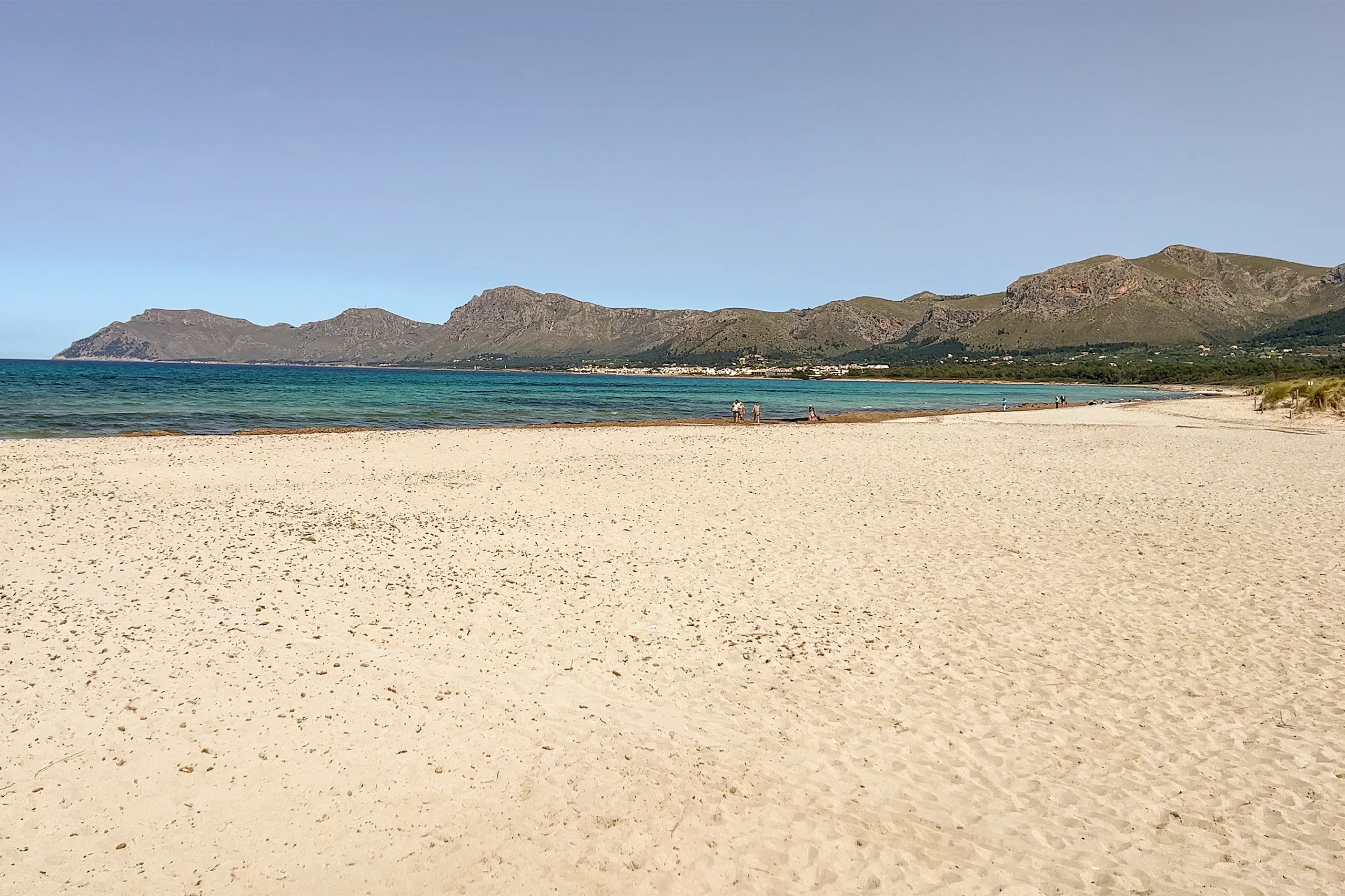 Weiter heller Sandstrand von Son Serra de Marina mit türkisfarbenem Flachwasser, fast menschenleer, im Hintergrund die Bergkette des Massís d'Artà unter blauem Himmel.
