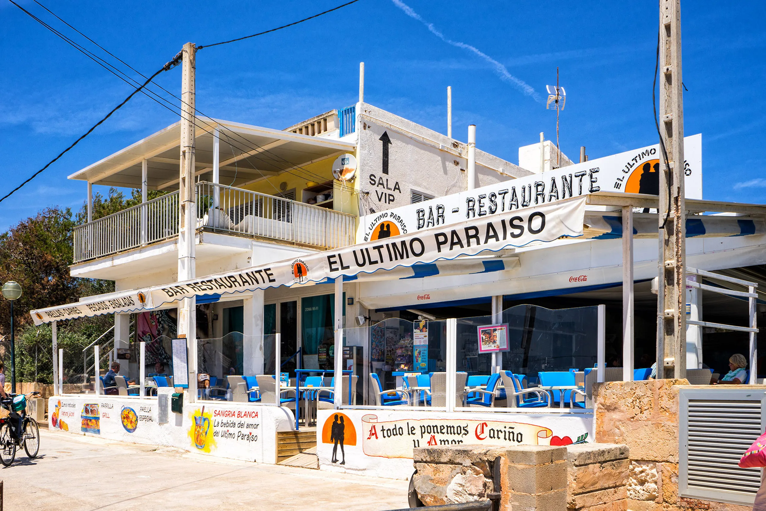 Zweigeschossiges weißes Strandlokal in Ses Covetes mit Terrasse, blauen Stühlen und großen handgemalten Schildern unter blauem Himmel.
