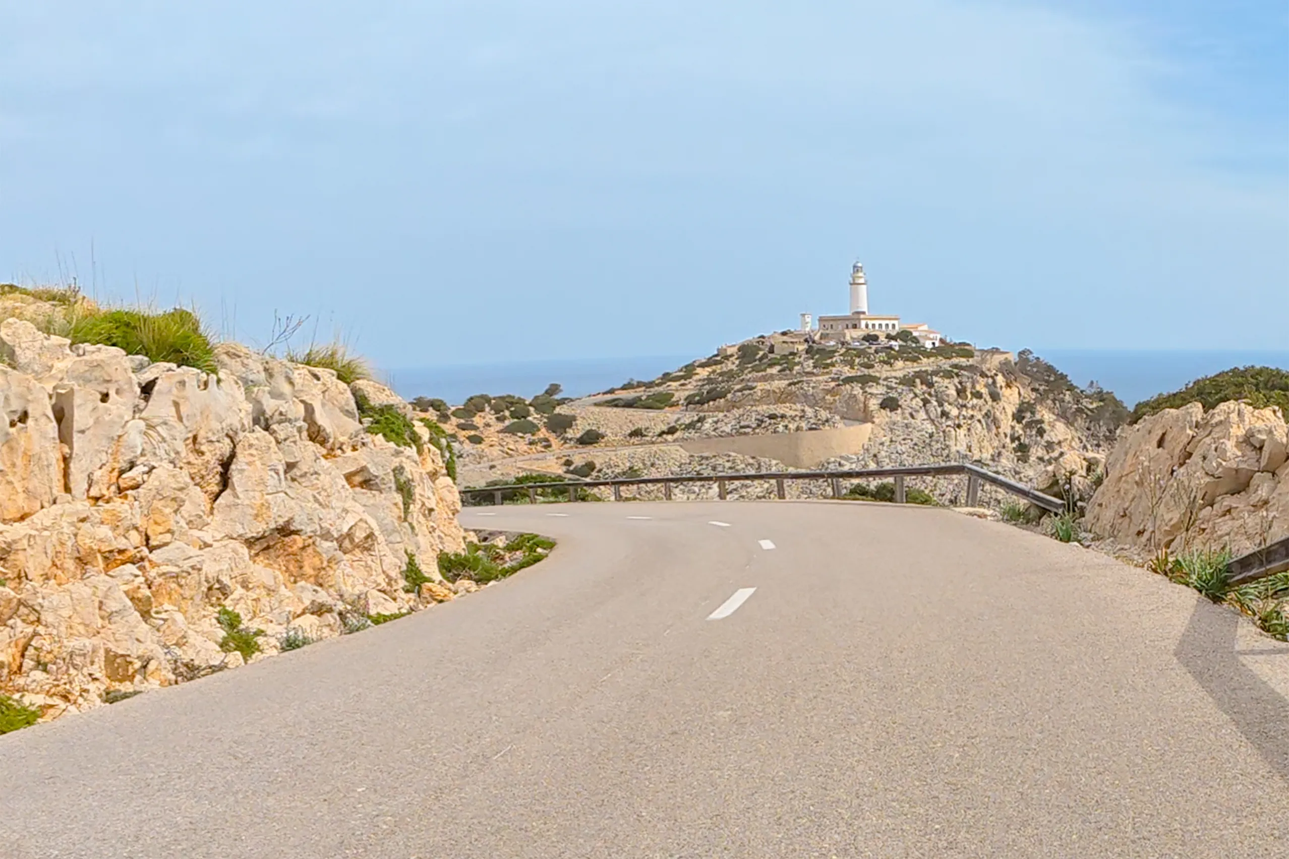 Die Straße zum Cap de Formentor führt zwischen hellen Felswänden hindurch, am Horizont steht der weiße Leuchtturm auf einer kahlen Kuppe über dem Meer.
