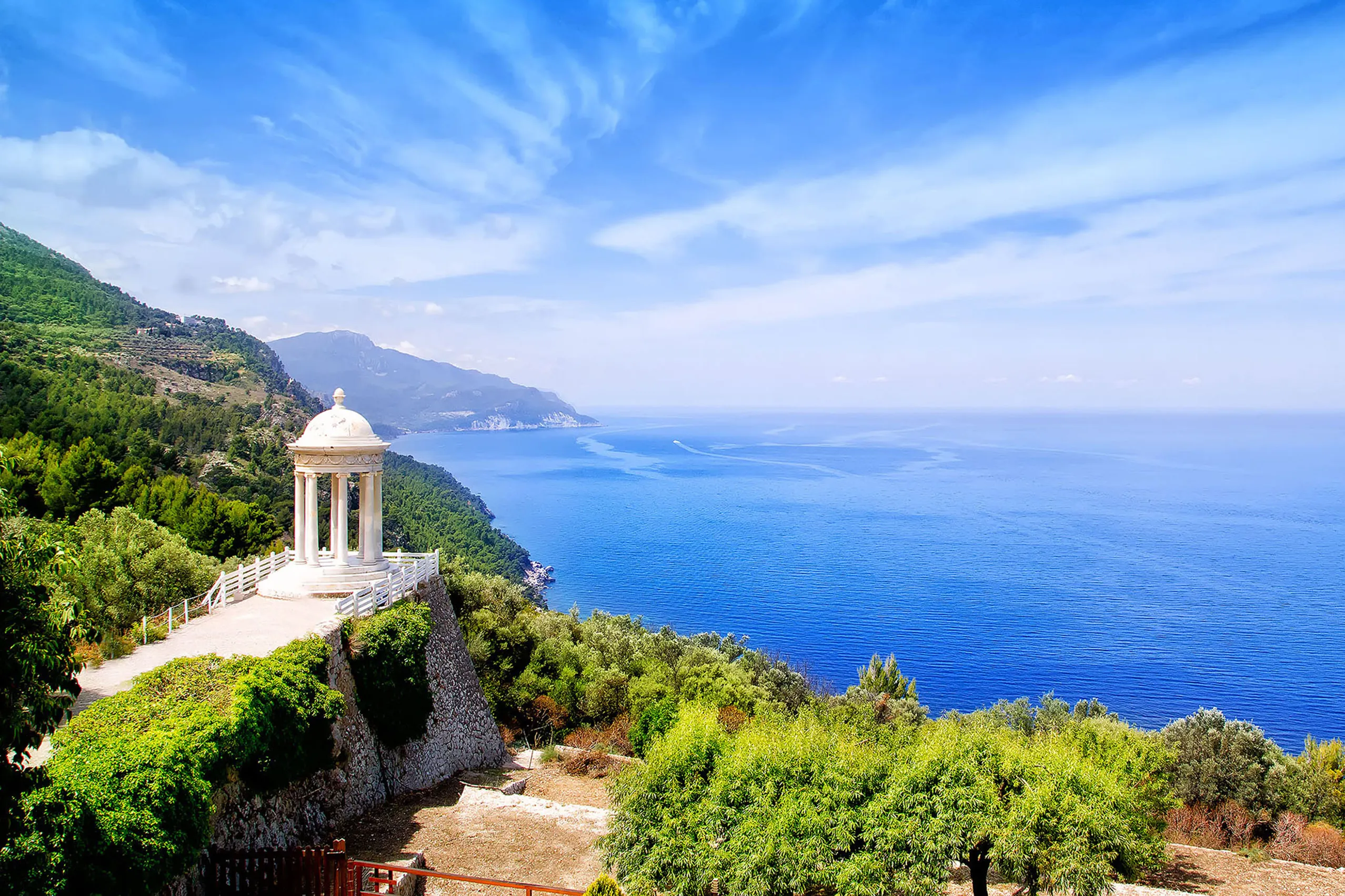 Weißer Rundtempel mit Säulen und Kuppel auf einer Terrasse hoch über dem tiefblauen Meer bei Son Marroig, dahinter die Steilküste der Tramuntana.

