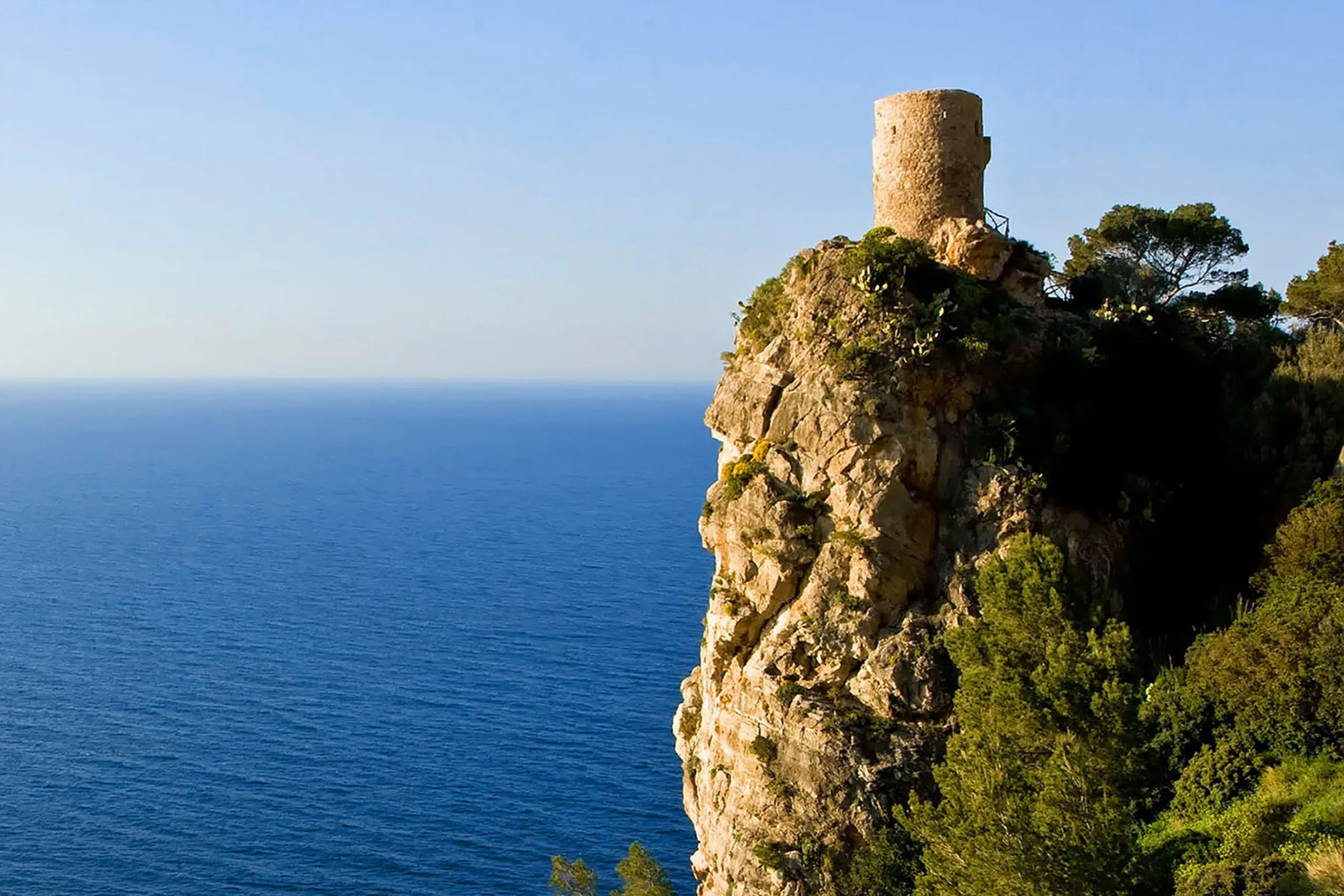 Runder steinerner Wachturm auf einem hohen, bewachsenen Felskopf über dem tiefblauen Meer, die Küste im Dunst dahinter.
